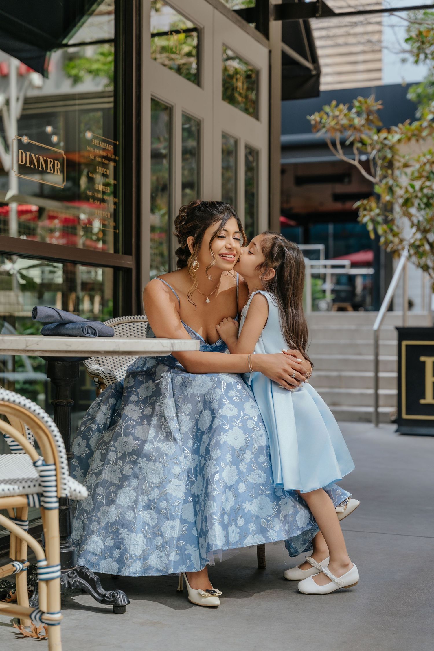 A woman in a blue floral dress sits outdoors as a young girl in a matching blue dress gives her a sweet kiss on the cheek.