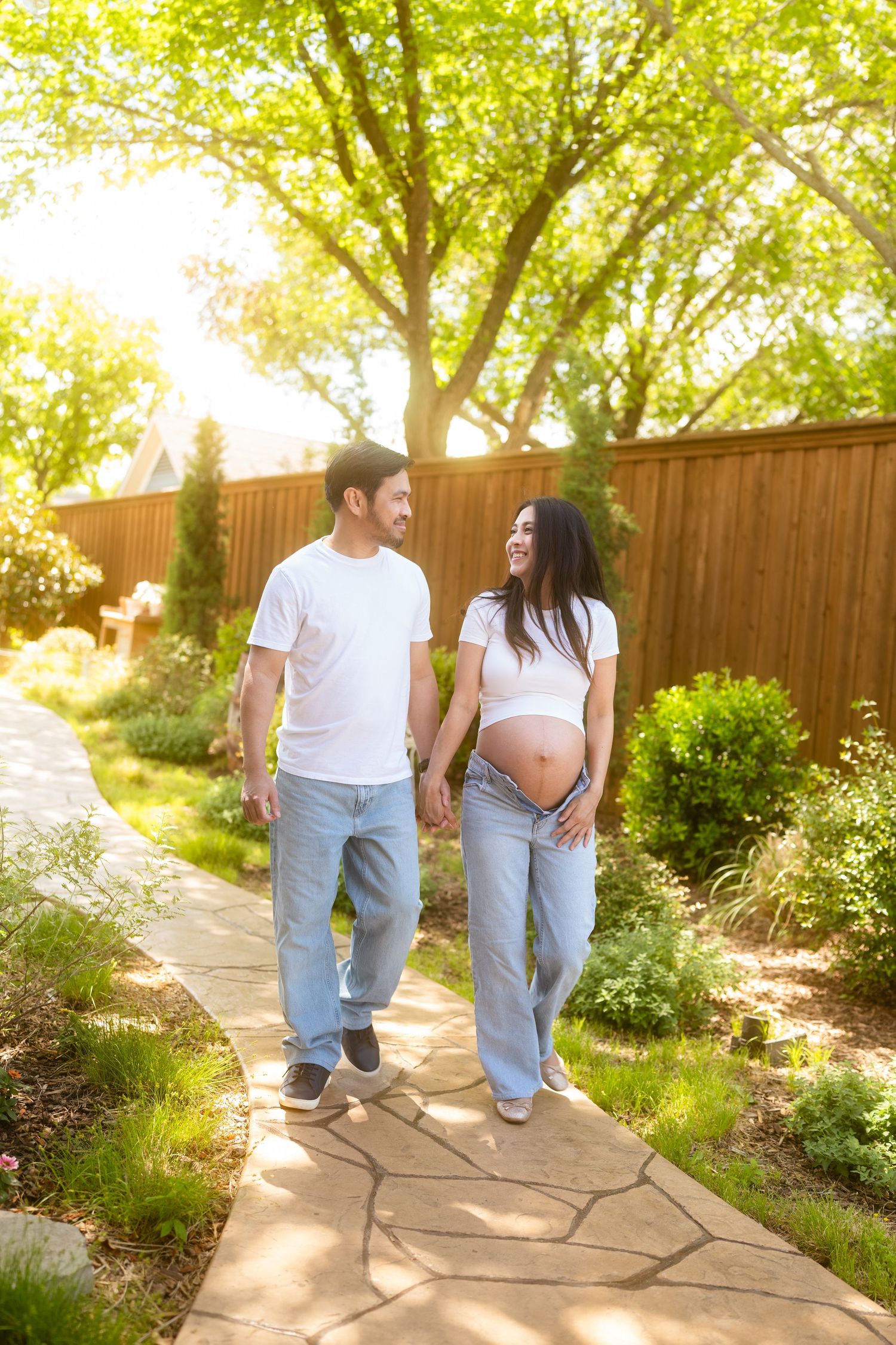Pregnant couple holding hands walking on garden path, wearing white shirts and jeans, surrounded by lush greenery.