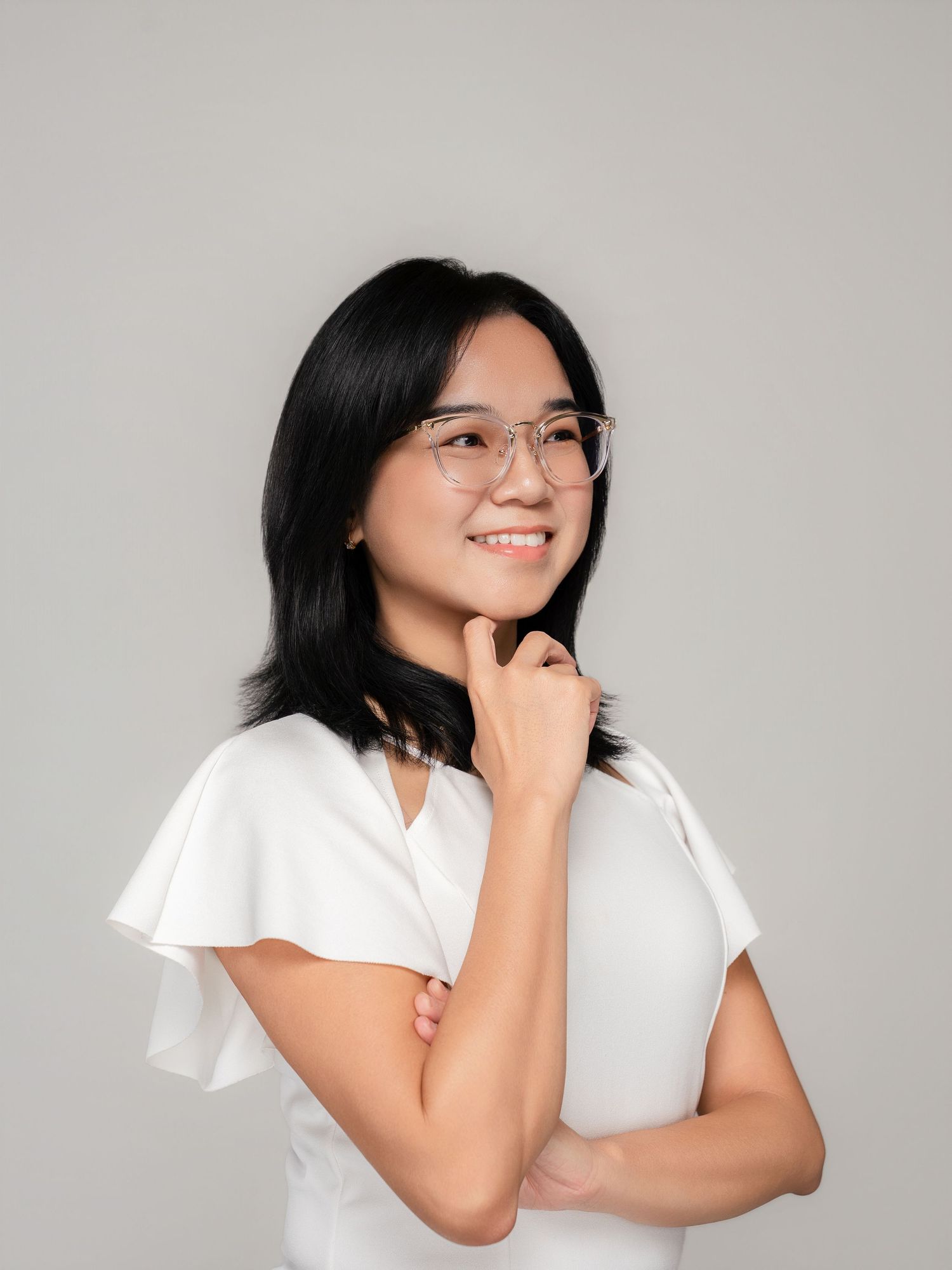 Young Asian woman in white ruffled top and clear glasses smiling confidently against a gray background.