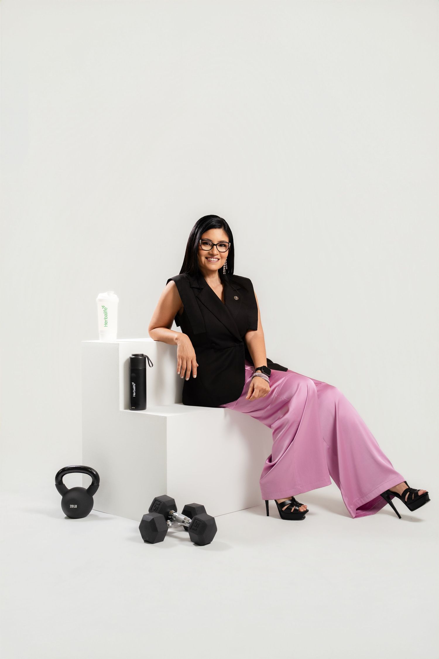 Woman in black top and pink pants sits on white cube with kettlebell and dumbbells nearby in a studio setting.