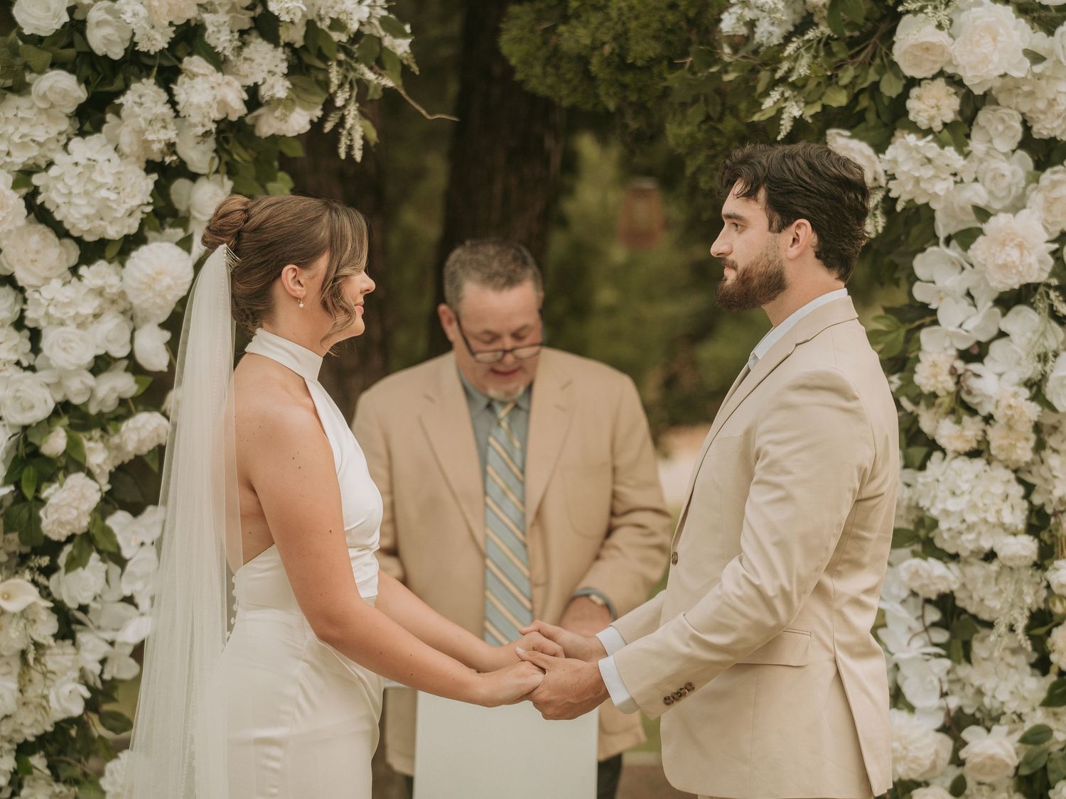 Bride and groom hold hands at outdoor wedding ceremony beneath white floral arch with officiant.