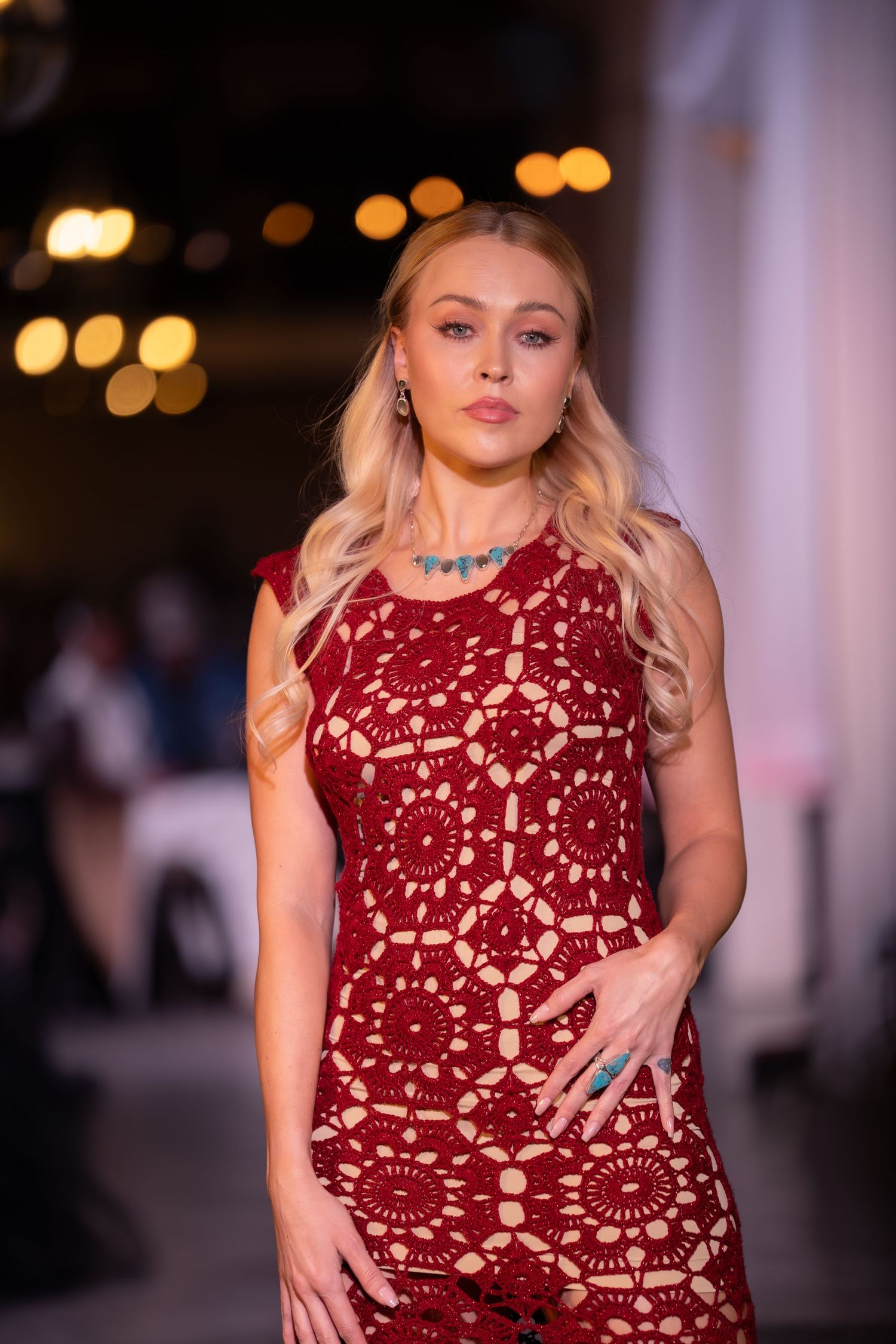 Model walks runway in burgundy lace cocktail dress with sleeveless design and fitted silhouette during fashion show.