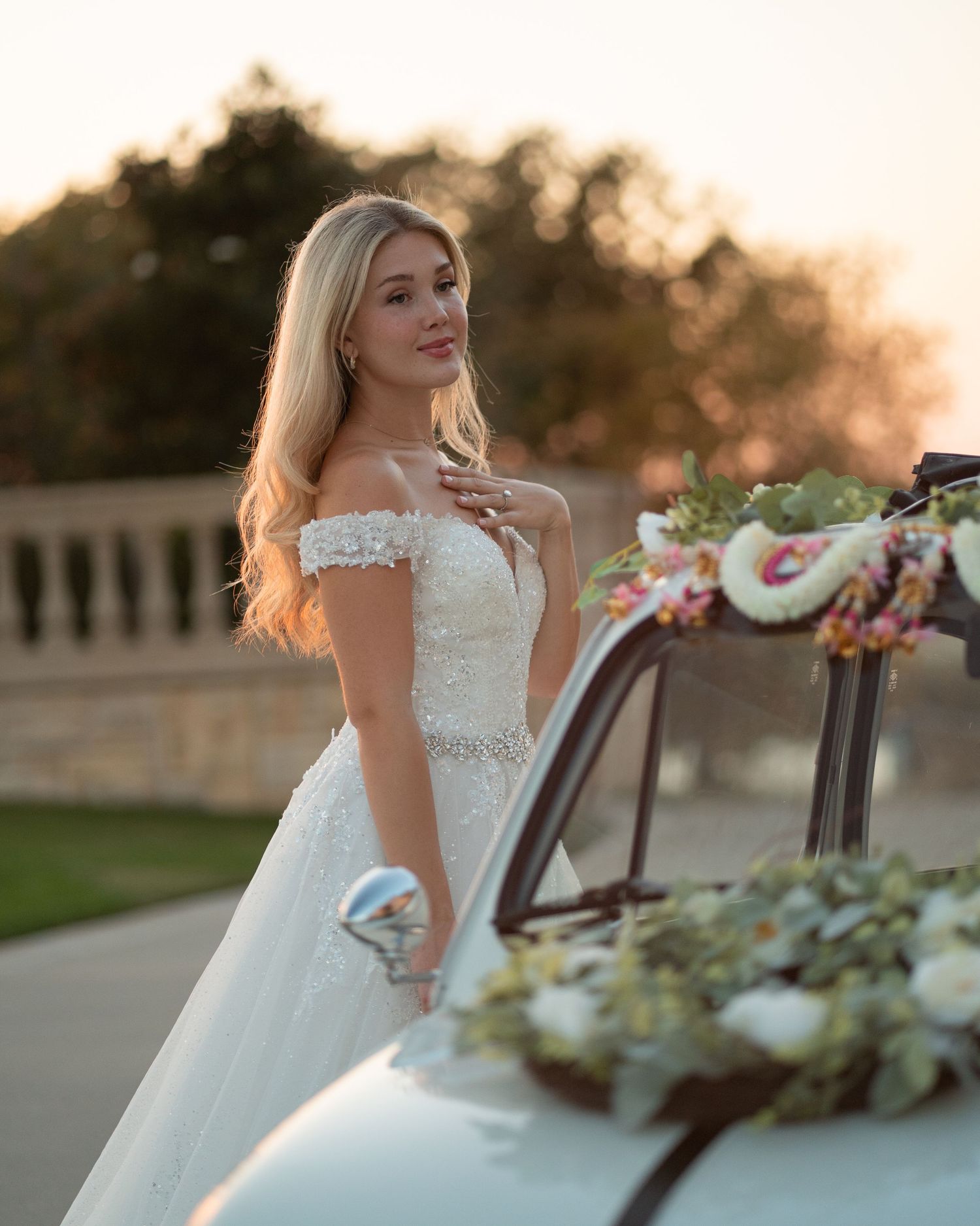 Bride in off-shoulder wedding gown posing near vintage furniture at sunset.