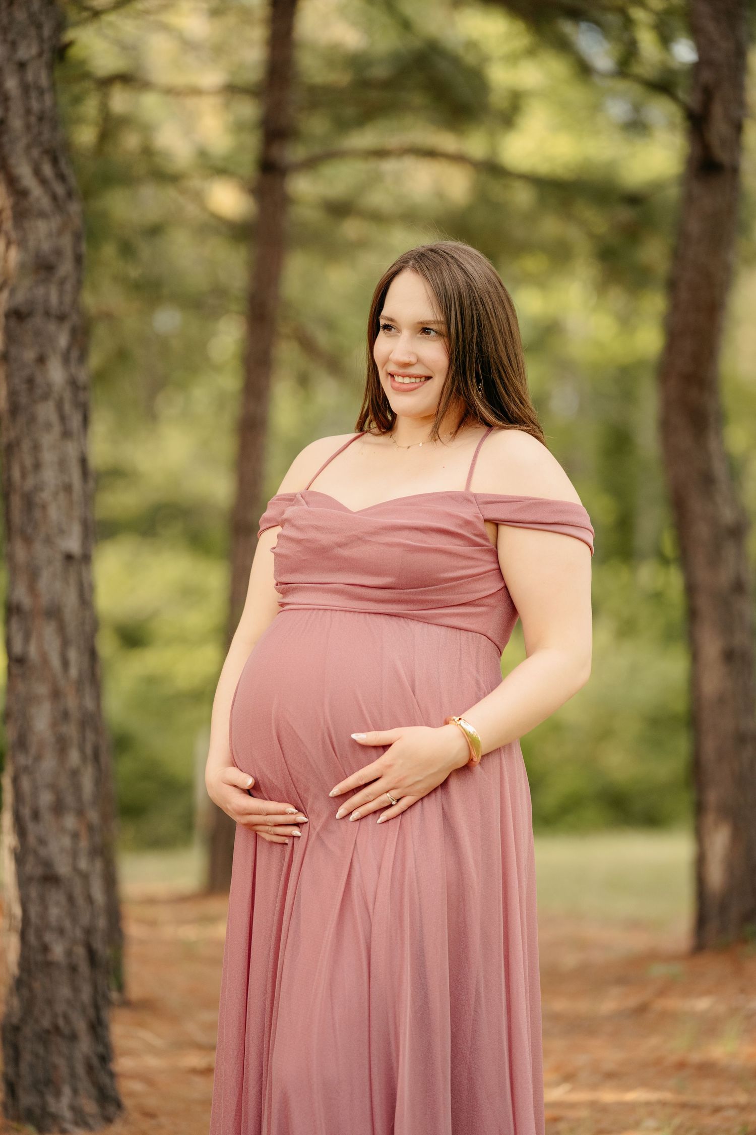 Smiling pregnant woman in a dusty rose off-shoulder gown cradling her baby bump outdoors among tall pine trees.