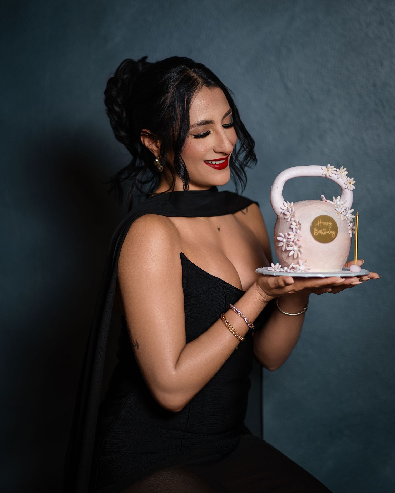 Woman in black dress holding elegant white floral birthday cake, smiling, dark background.