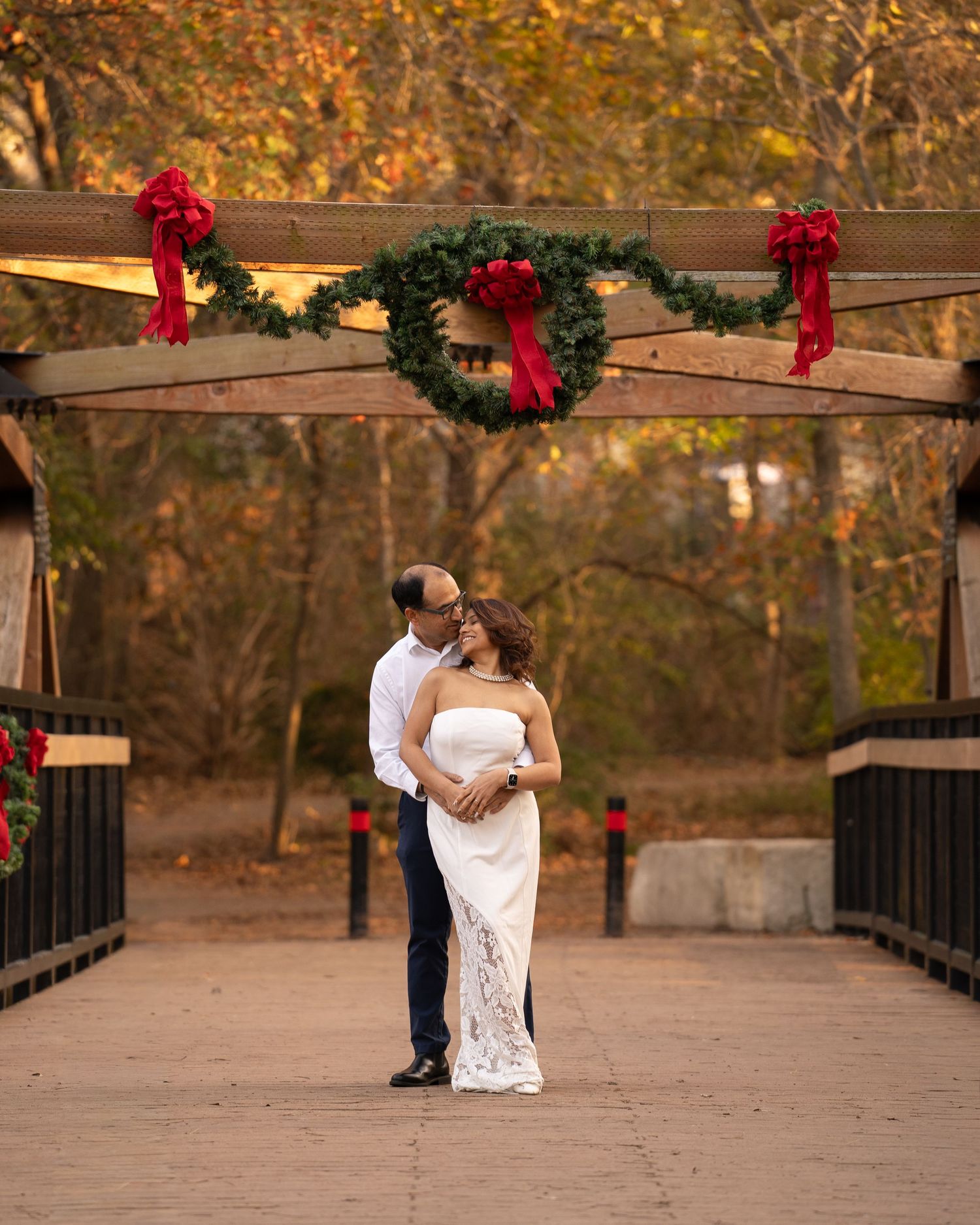 Couple embraces on a decorated bridge with Christmas garland and red bows, surrounded by autumn foliage.