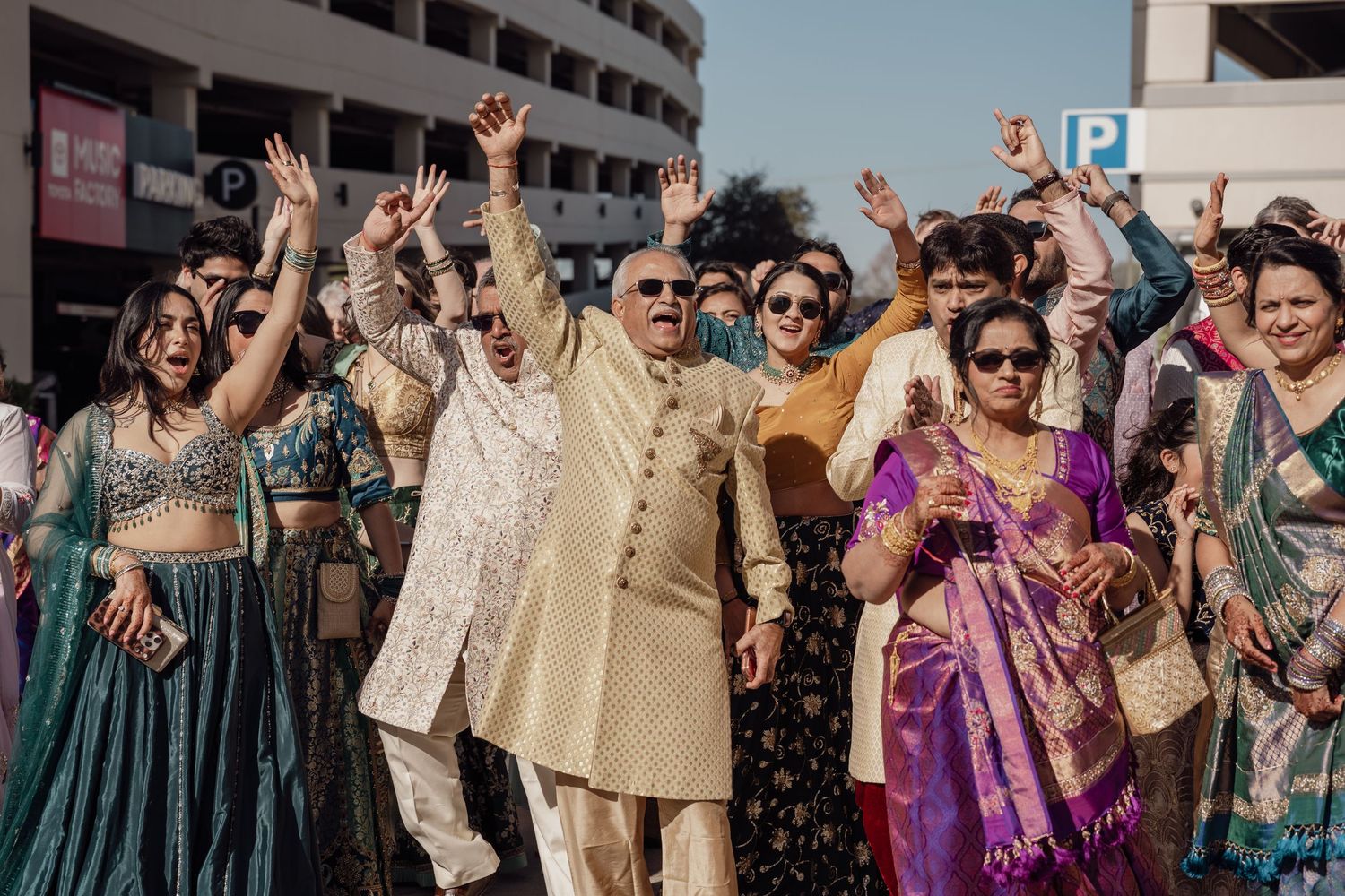 Joyful Indian wedding guests in vibrant traditional attire celebrating outdoors with raised hands and smiling faces.