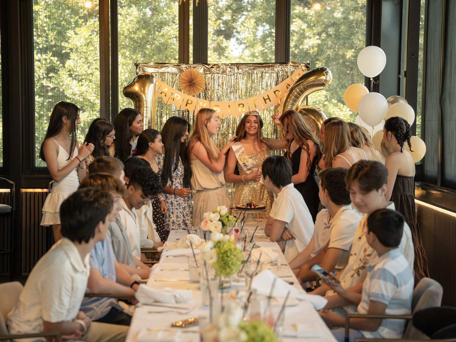 Elegant birthday party celebration with guests gathered around a long table, gold balloon decorations, and happy birthday banner.
