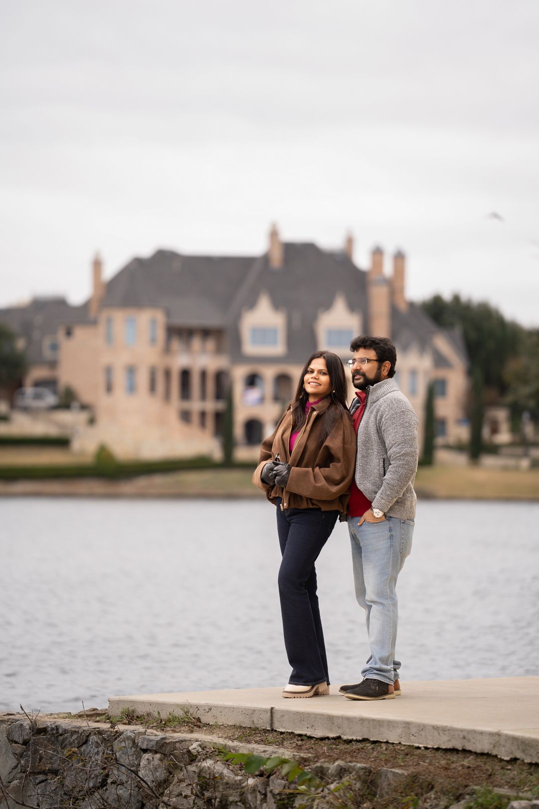 Couple posing by a lakeside with luxury homes in the background.