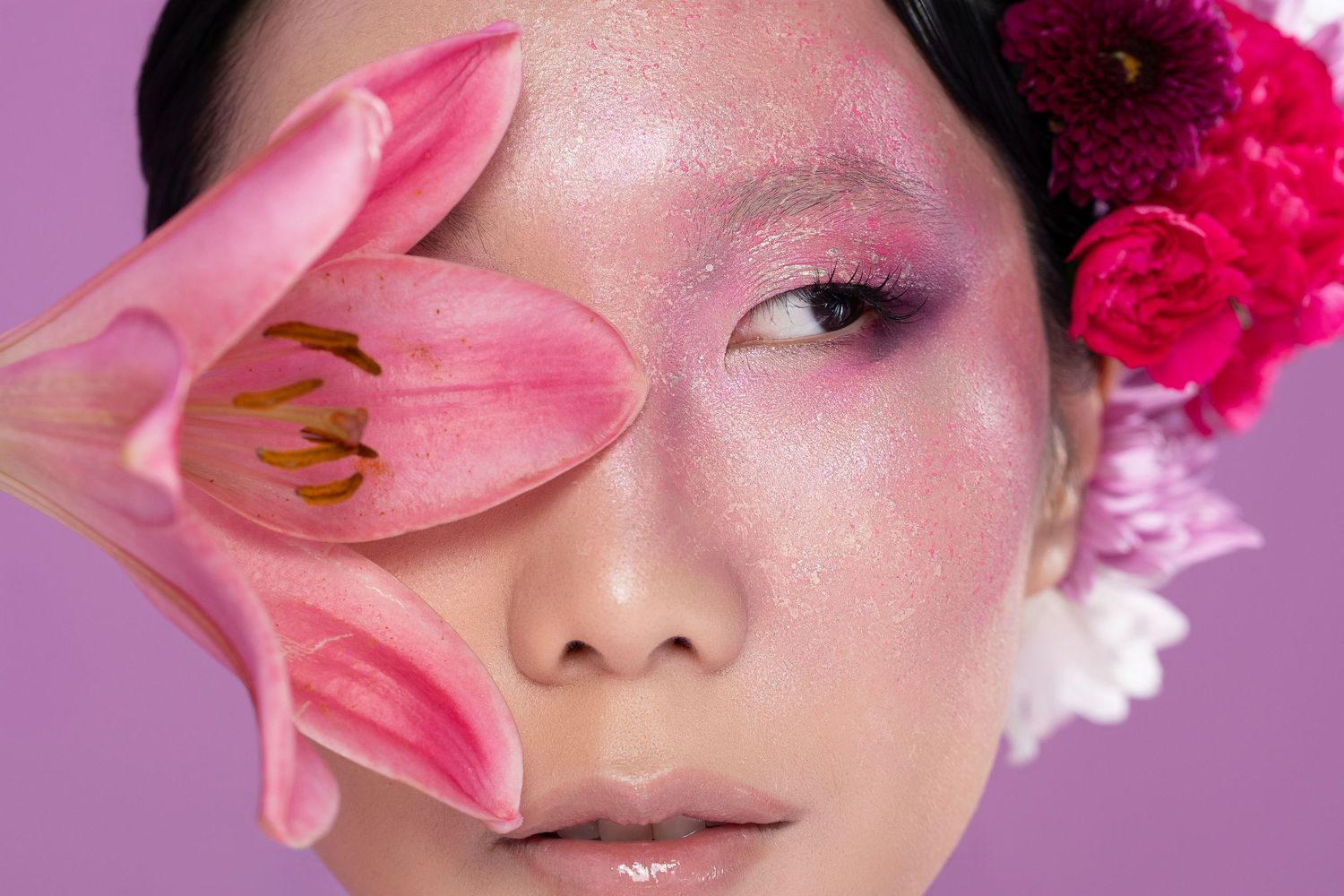Close-up of Asian woman with pink glitter eye makeup, holding a pink lily flower over one eye, with floral hair accessories.