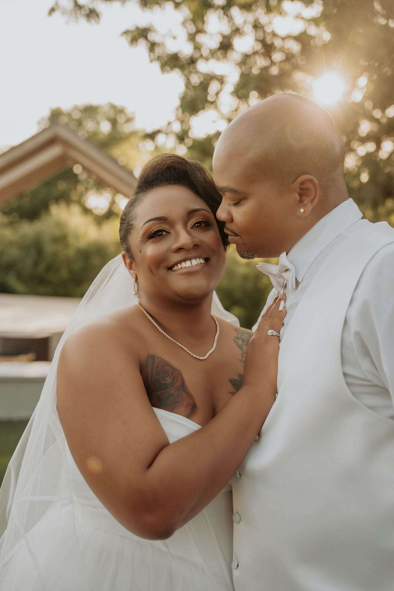 A bride smiles joyfully as her partner kisses her cheek during a golden hour outdoor wedding ceremony.