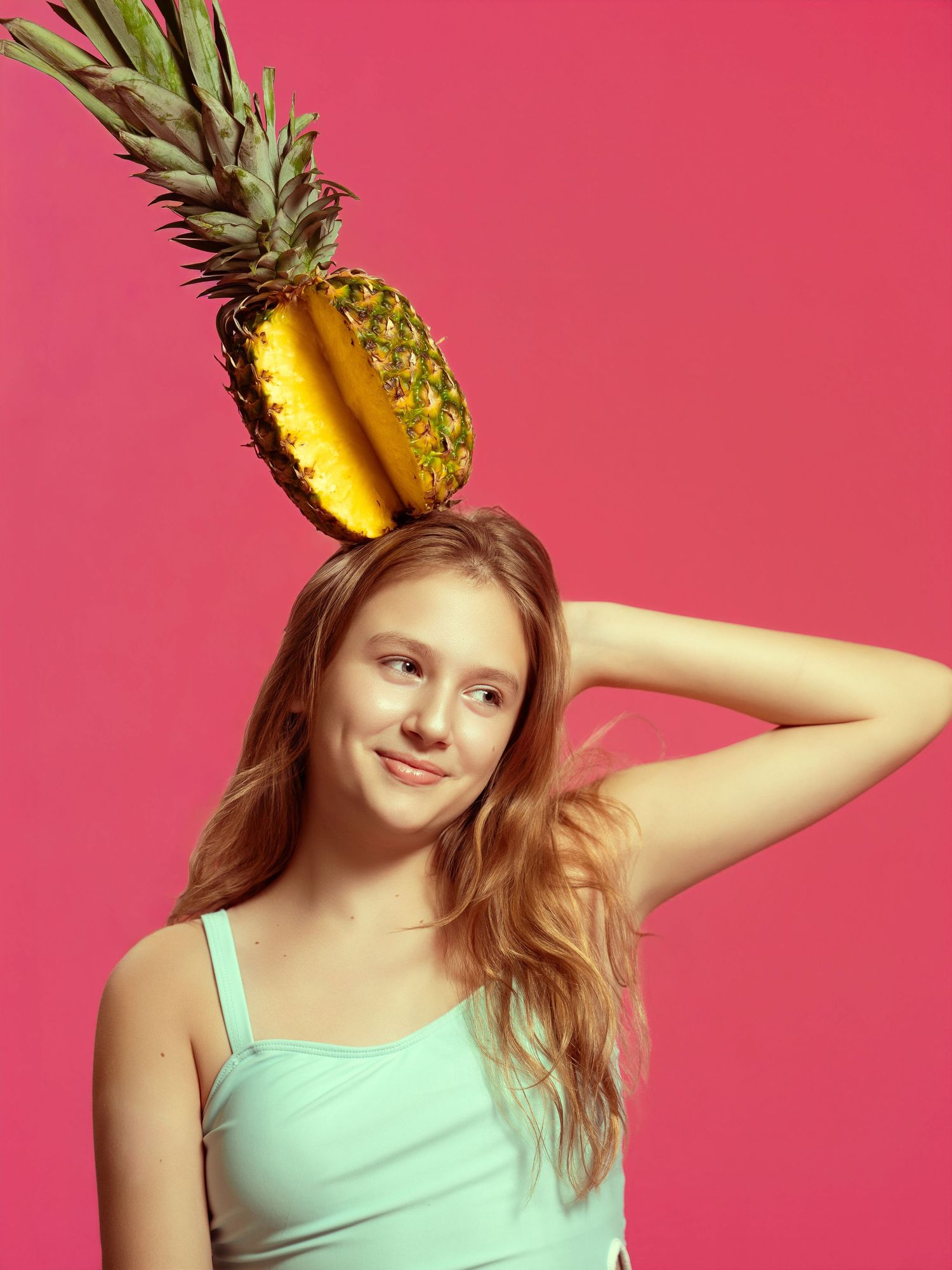 Young woman balancing a sliced pineapple on her head against a vibrant pink background.