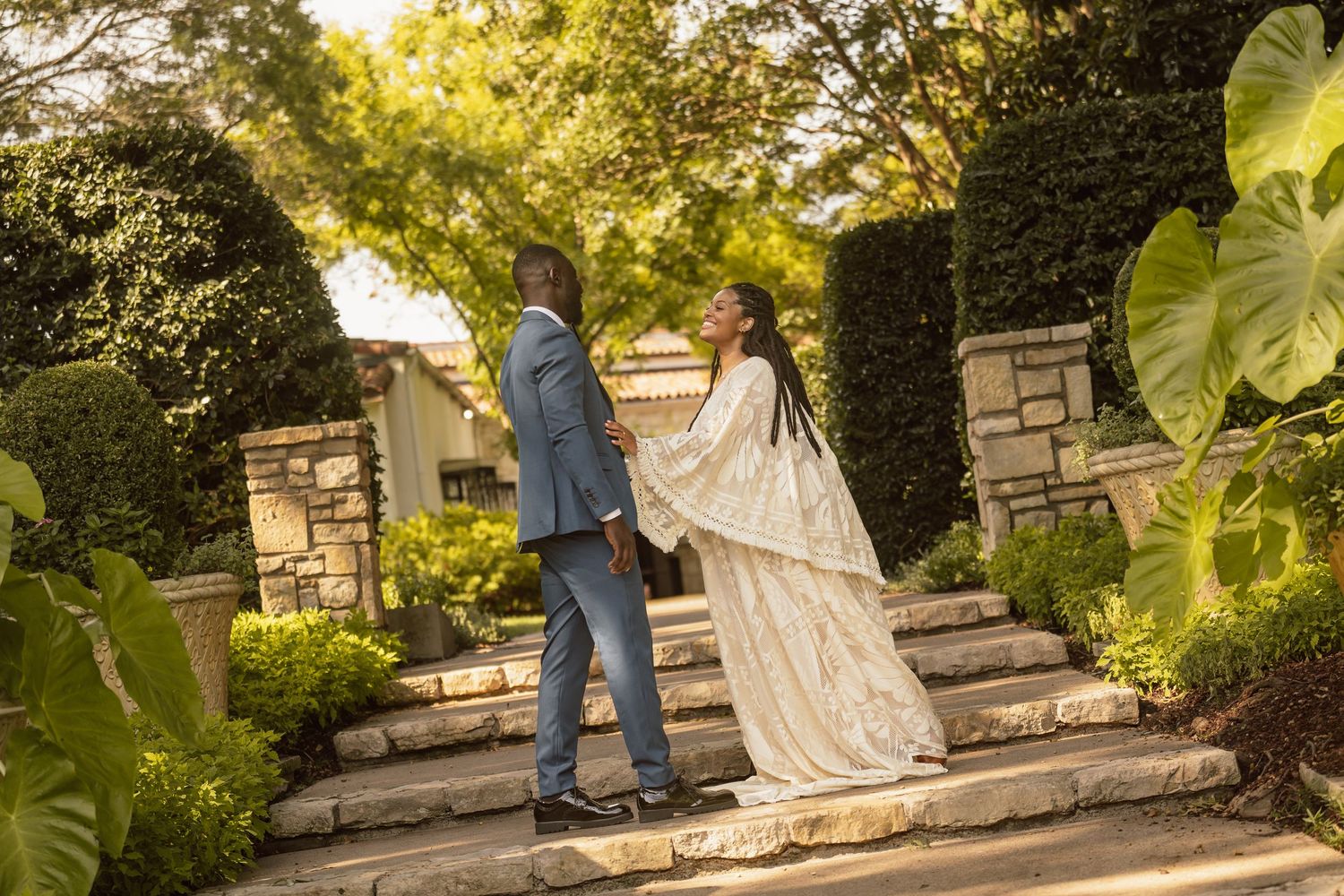 Couple in elegant wedding attire standing on stone steps surrounded by lush greenery in a garden setting.