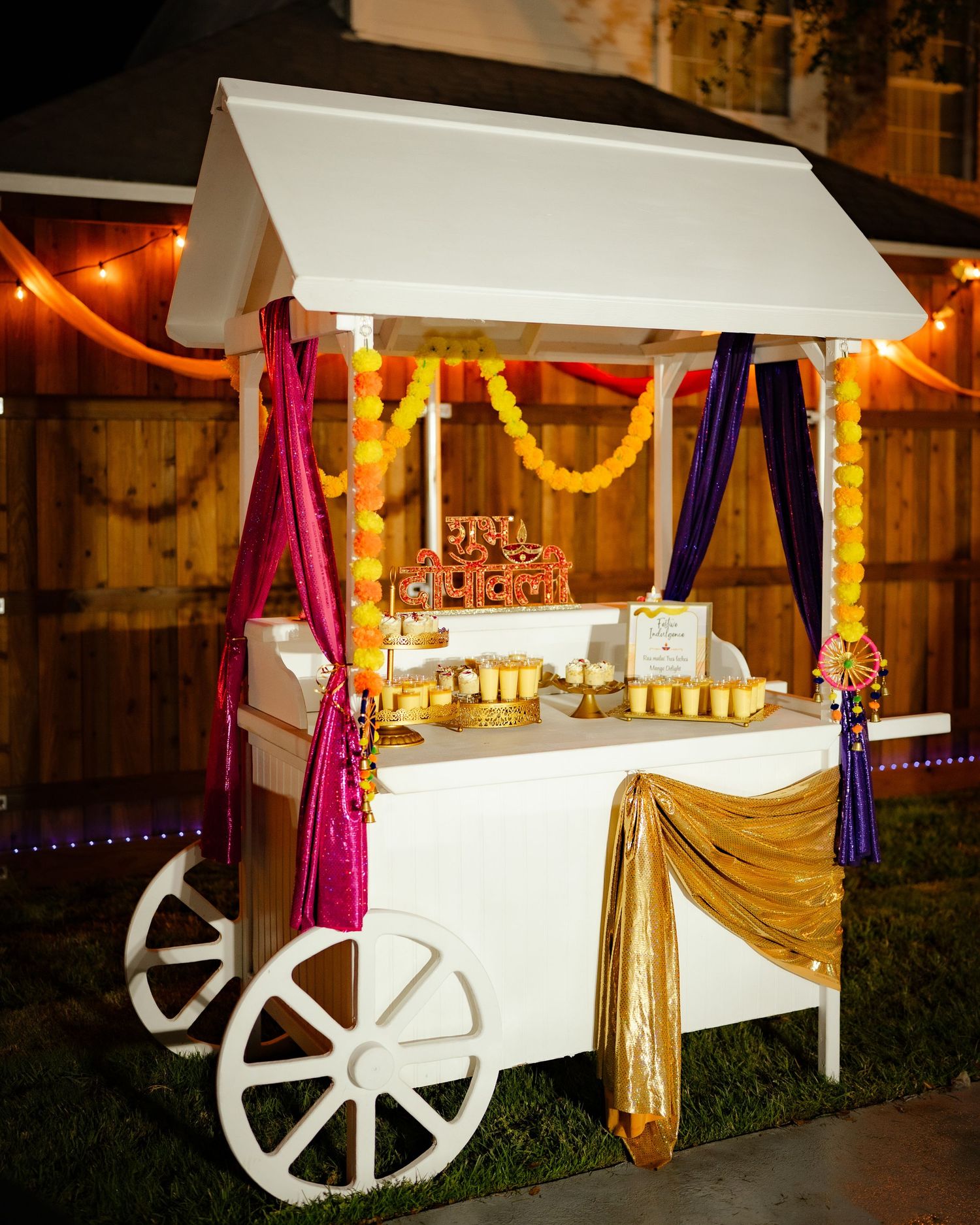 A white carnival food cart decorated with colorful drapes and string lights creates a festive atmosphere at an outdoor event.