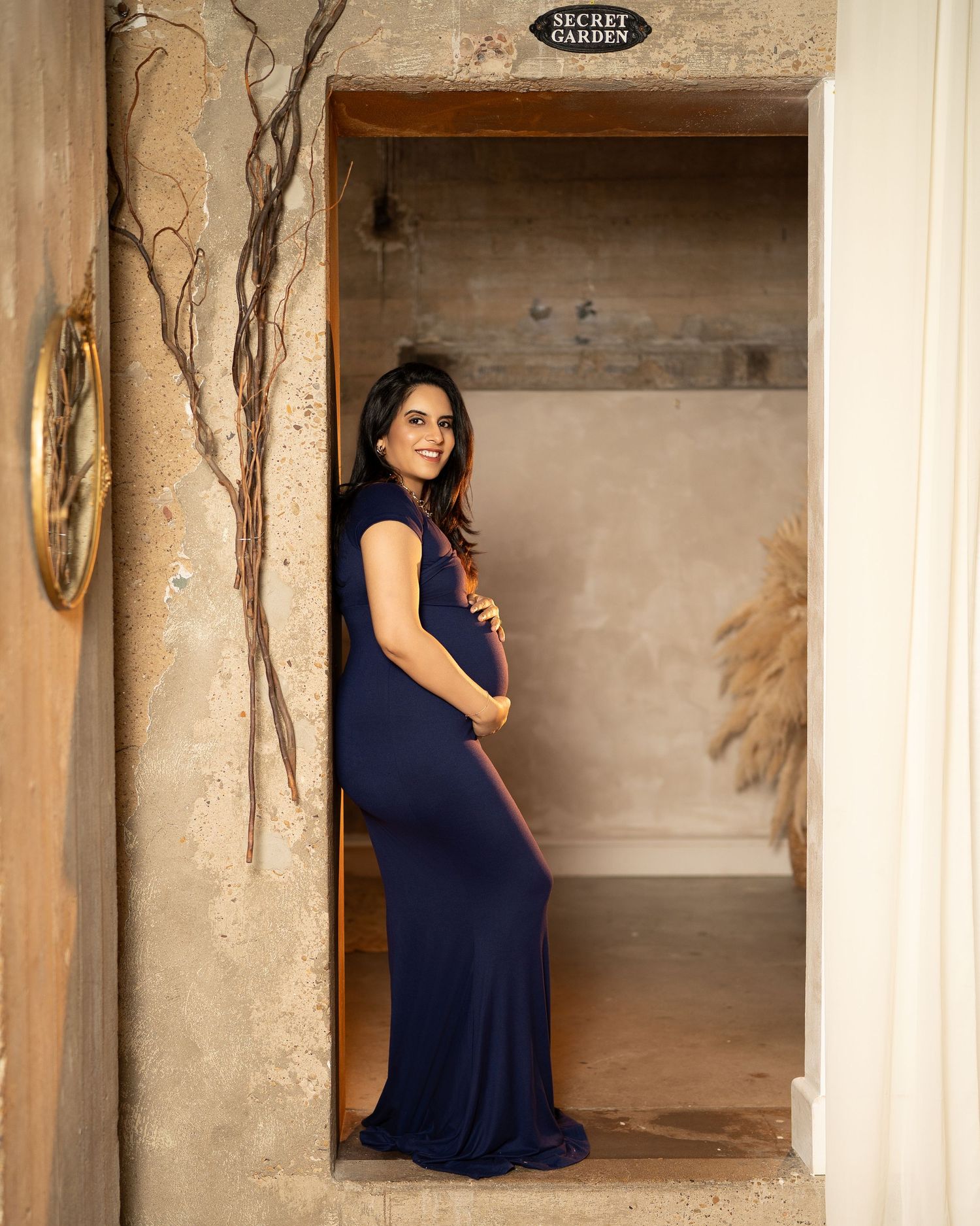Pregnant woman in navy blue maternity gown posing in a doorway labeled Secret Garden, smiling gracefully.