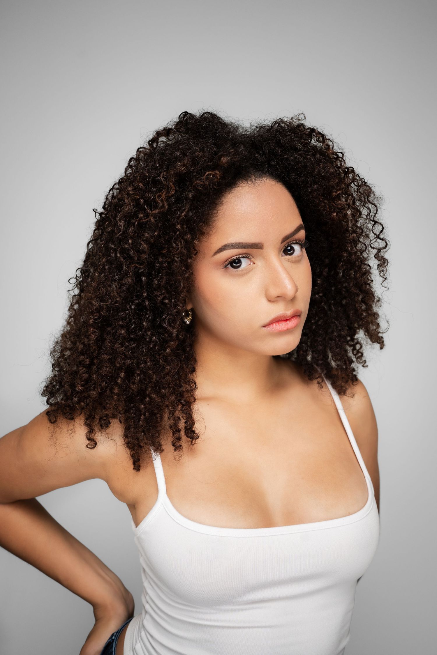 Young woman with voluminous curly dark hair wearing a white tank top, posing against a gray background.