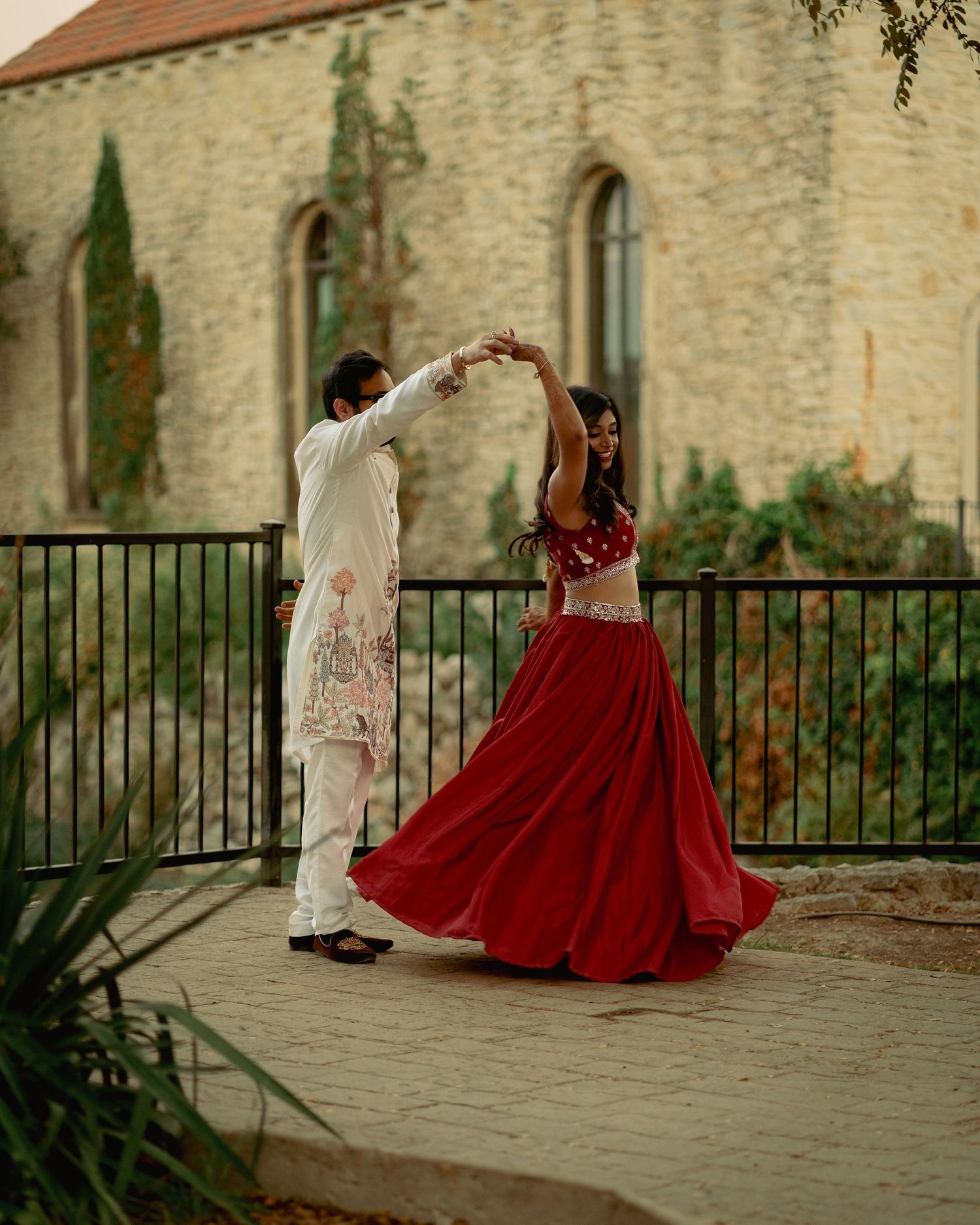 Couple dancing outdoors in traditional Indian attire, woman in red lehenga, man in white sherwani, near stone building.