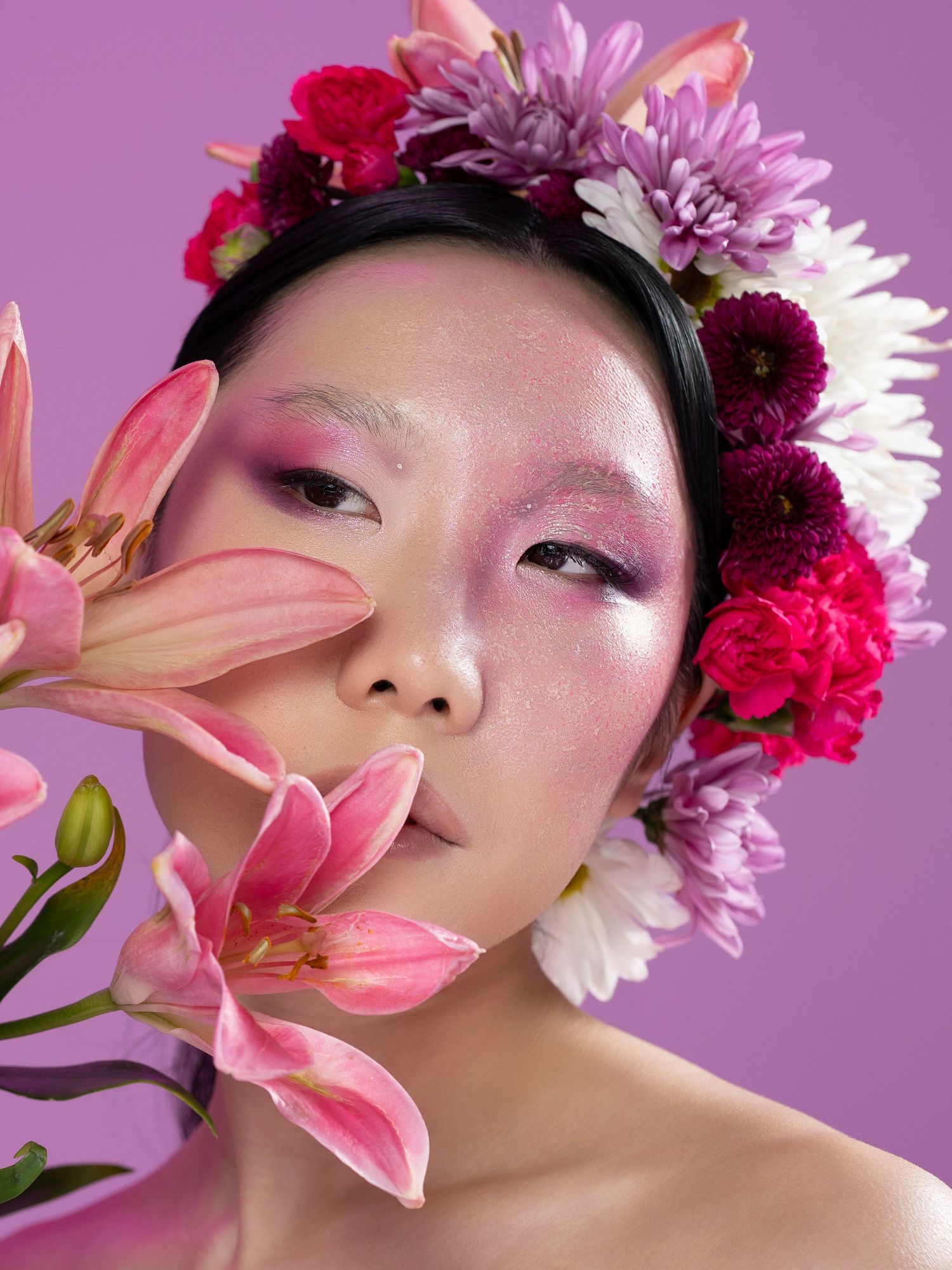 A woman with pink floral crown and matching makeup holds pink lilies against a purple background.