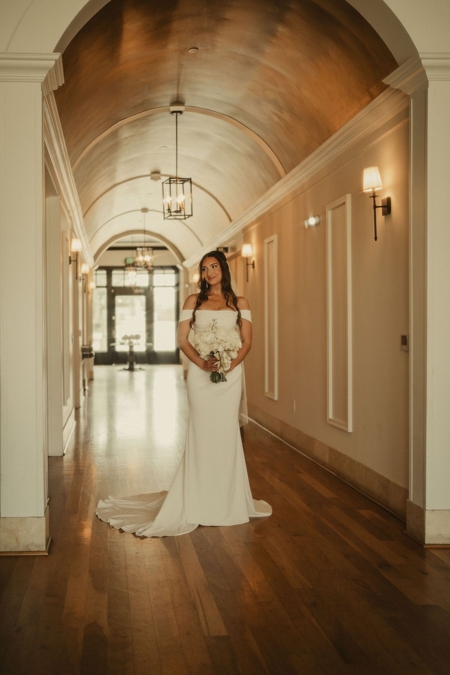 Bride in off-shoulder white gown holding bouquet stands in elegant arched hallway with warm lighting and hardwood floors.