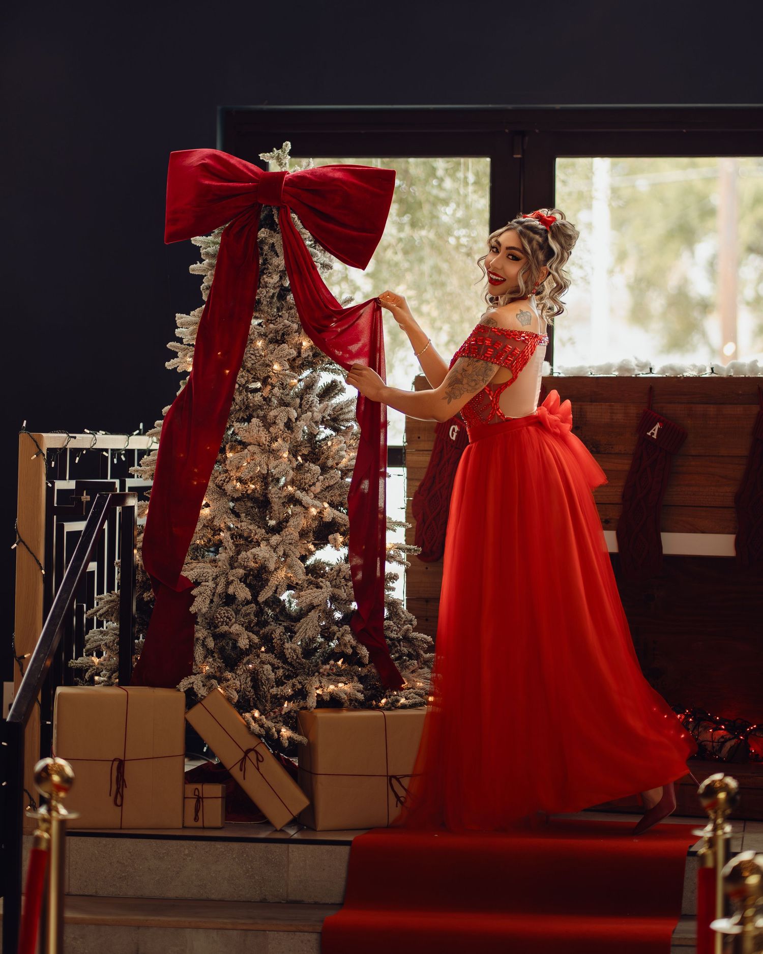 Woman in red ball gown arranging a large red bow on a decorated Christmas tree with gifts below.