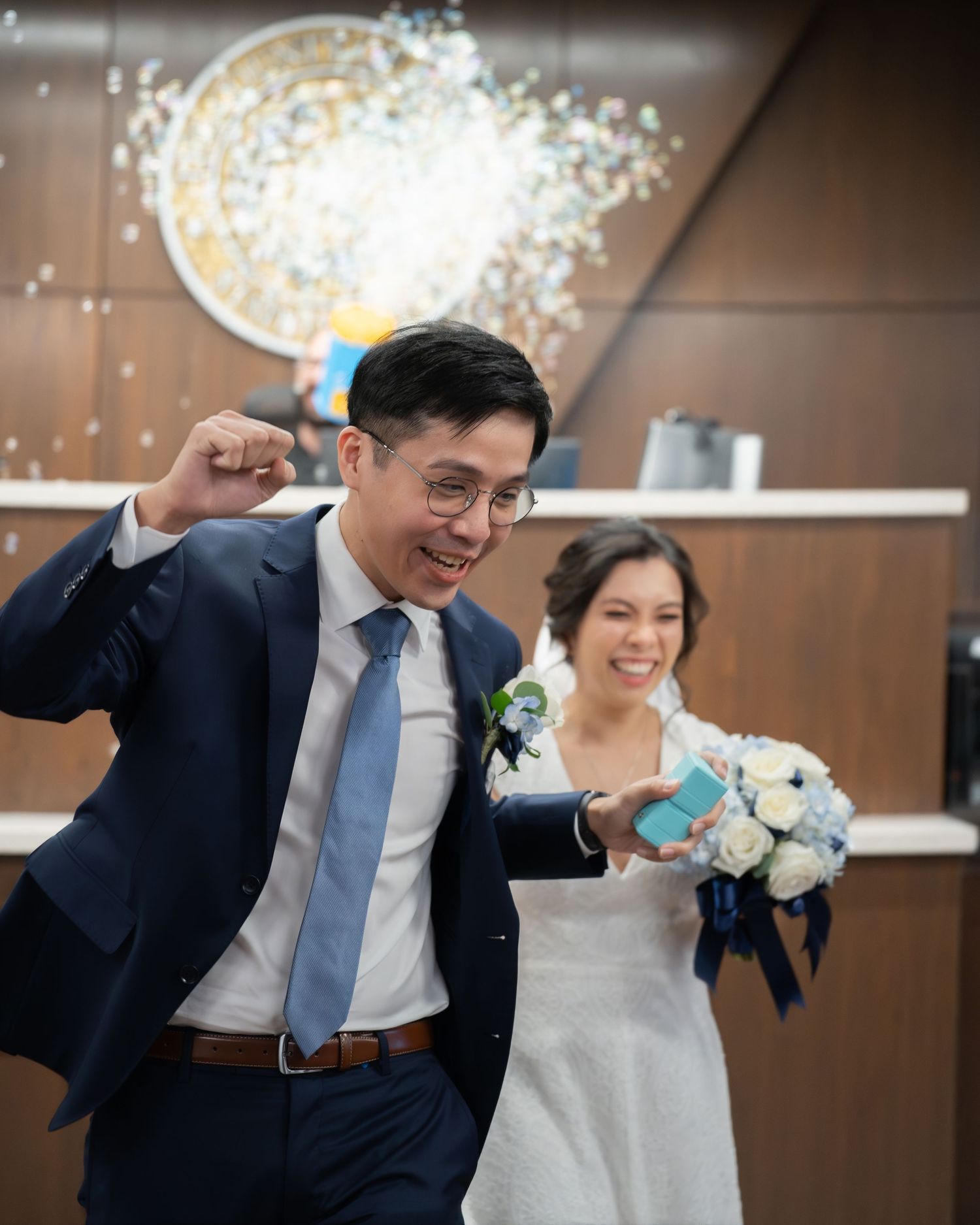 Joyful groom in navy suit pumps fist while bride in white dress holds bouquet as confetti falls at wedding ceremony.