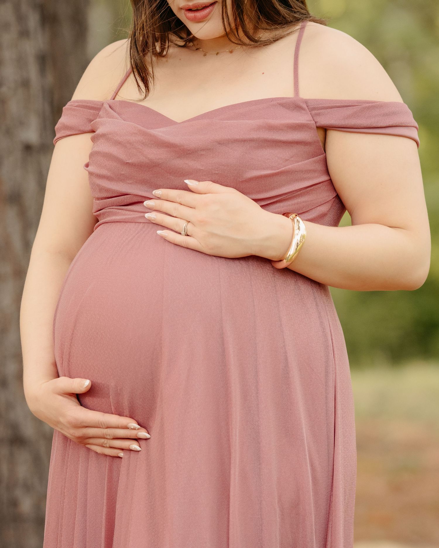 Pregnant woman in a dusty rose off-shoulder maternity gown cradling her baby bump outdoors.