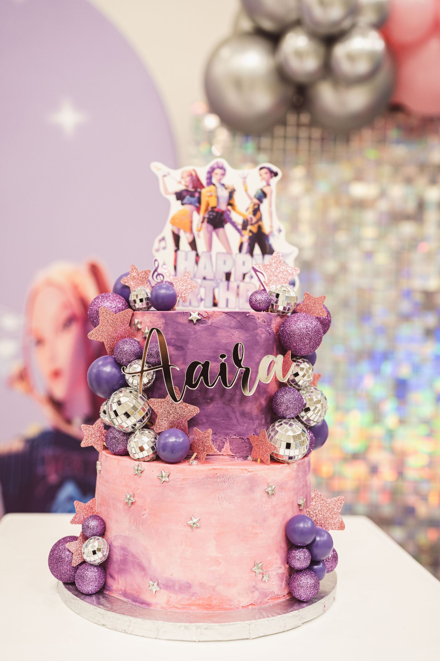 Pink and purple birthday cake decorated with sparkly ornaments, macarons and a custom name topper on a glittery background.