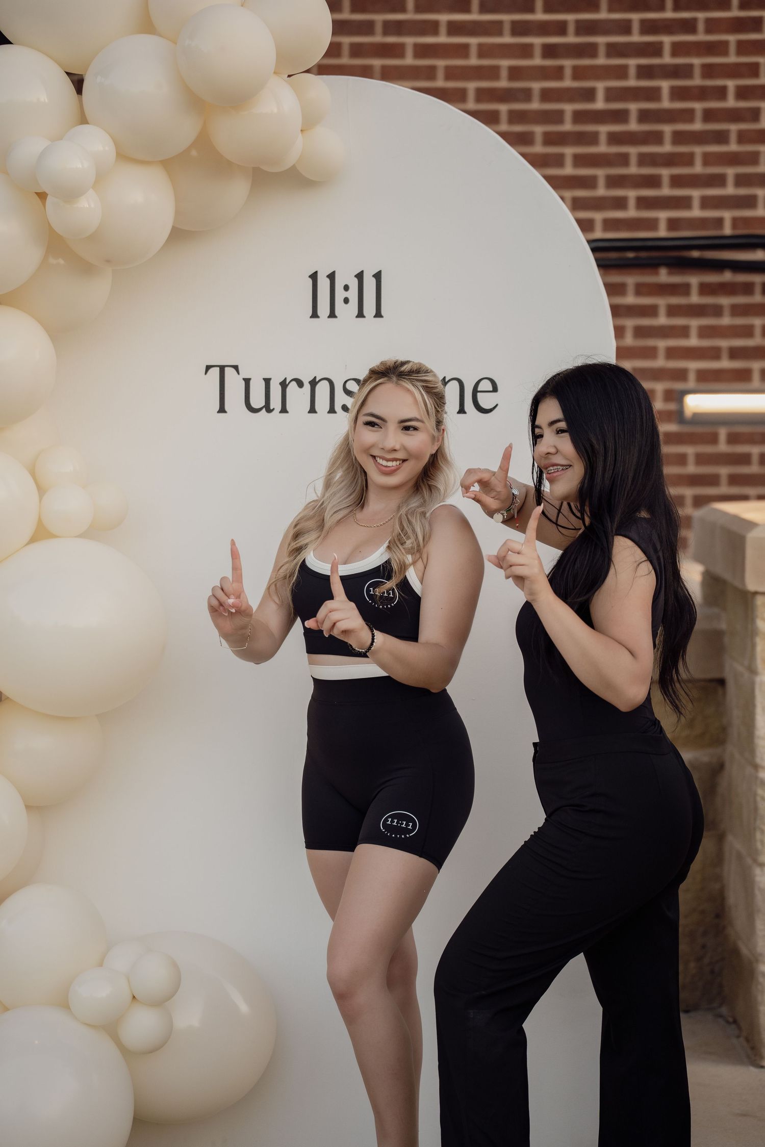 Two women pose playfully in front of a 11:11 Turnstone sign surrounded by cream balloons.