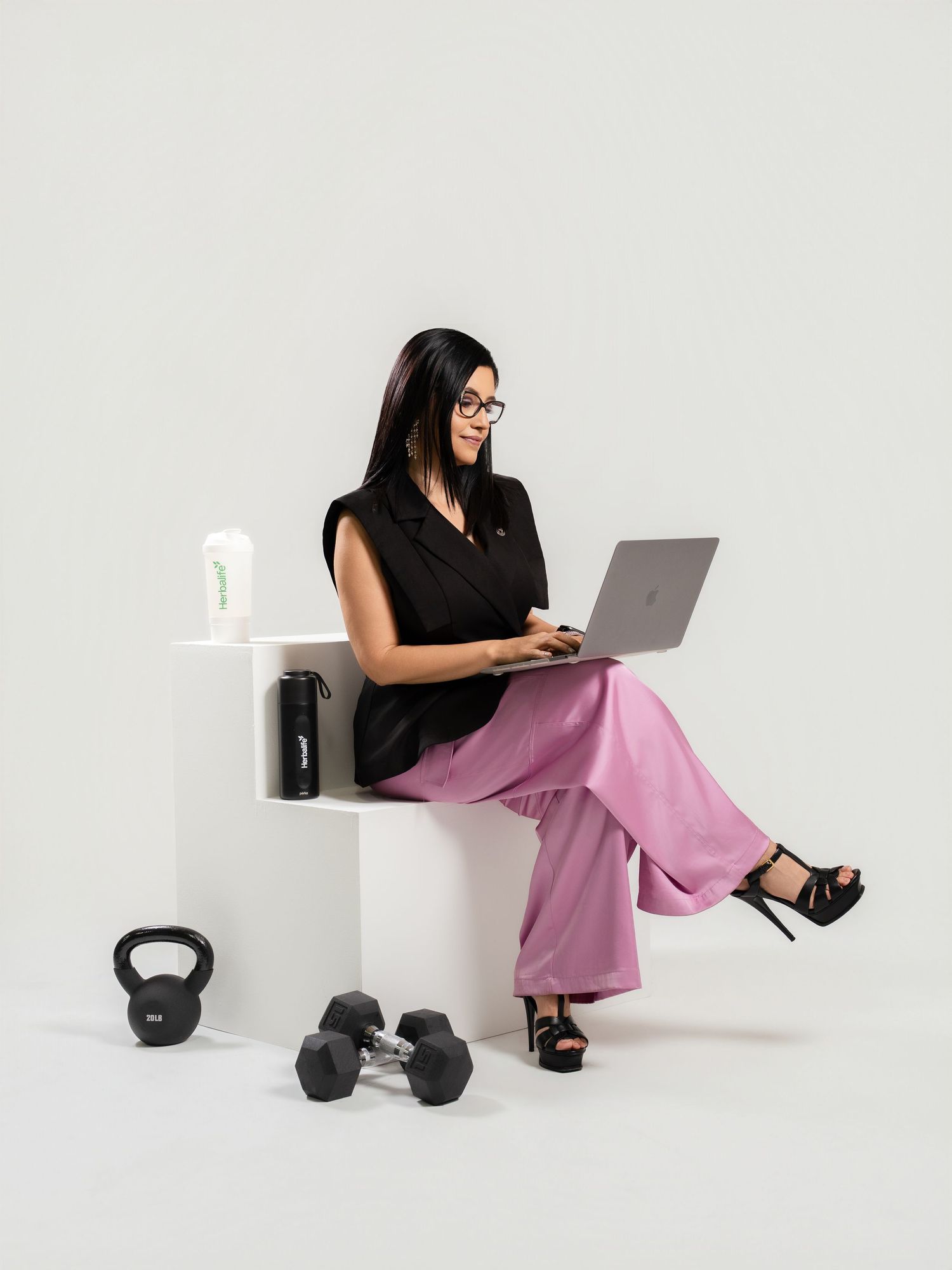 Woman in pink pants and black top sits on white cube with laptop, kettlebell and dumbbells nearby.