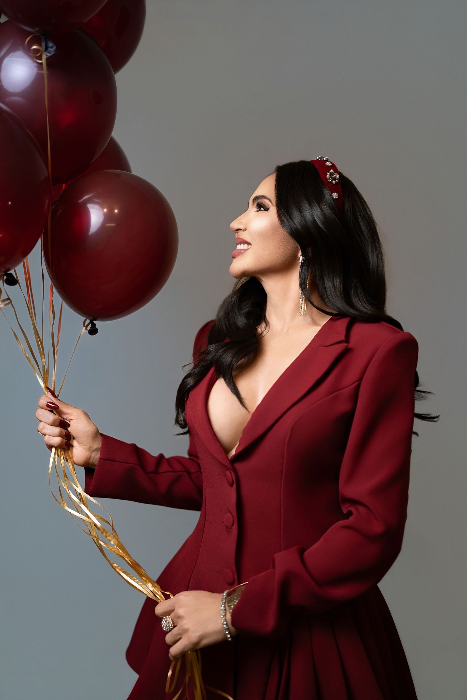 A smiling brunette woman in a burgundy suit holds matching dark red balloons against a gray background.