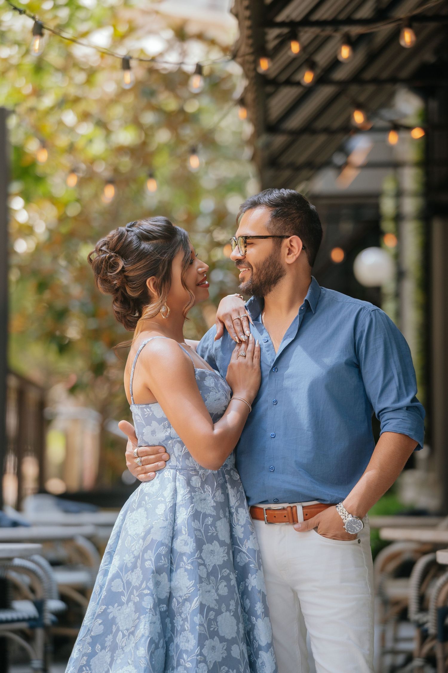 Couple in elegant attire embracing outdoors under string lights, woman in blue floral dress, man in blue shirt and white pants.