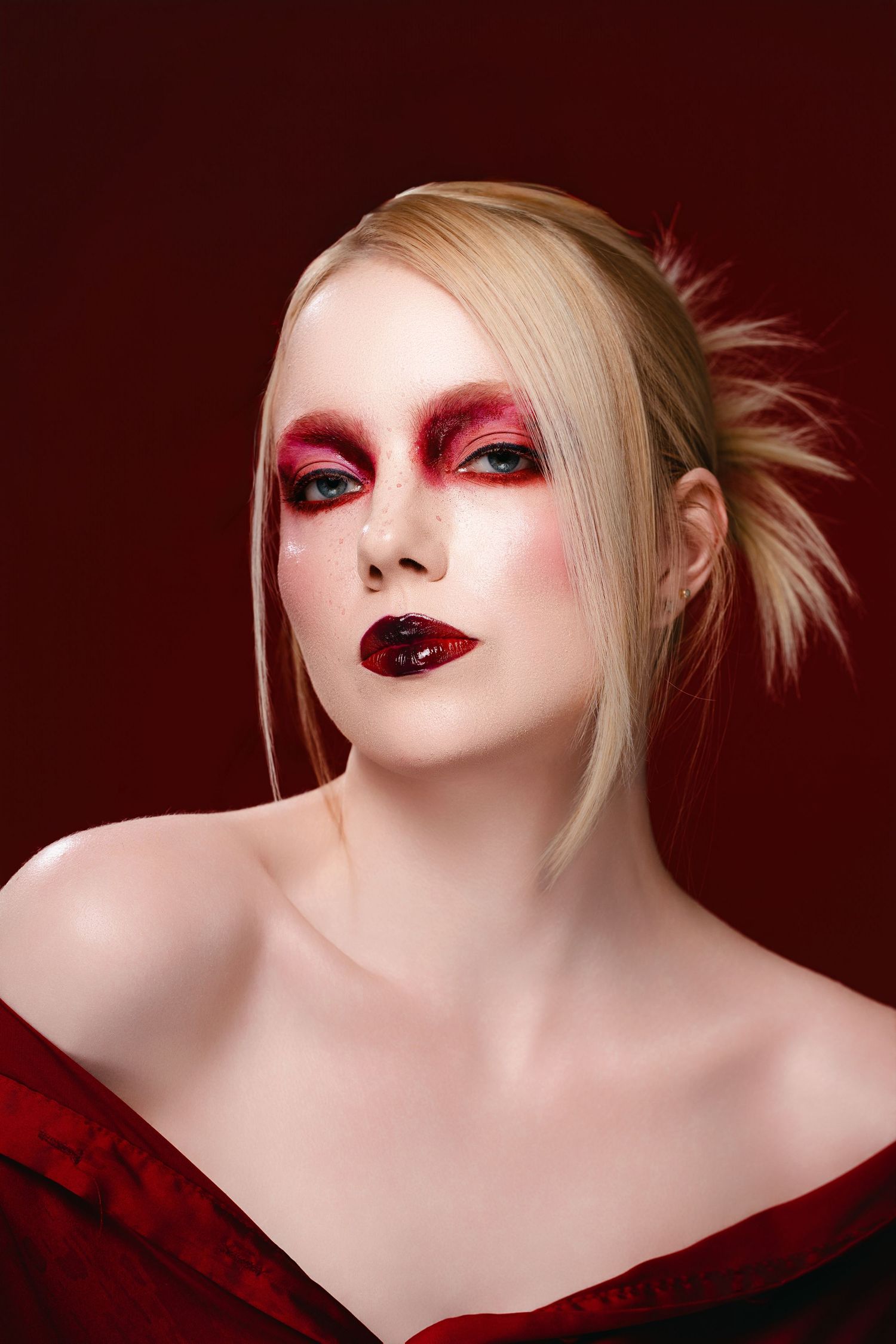 Blonde woman with dramatic red and dark lip makeup, wearing an off-shoulder red velvet top against a dark red background.