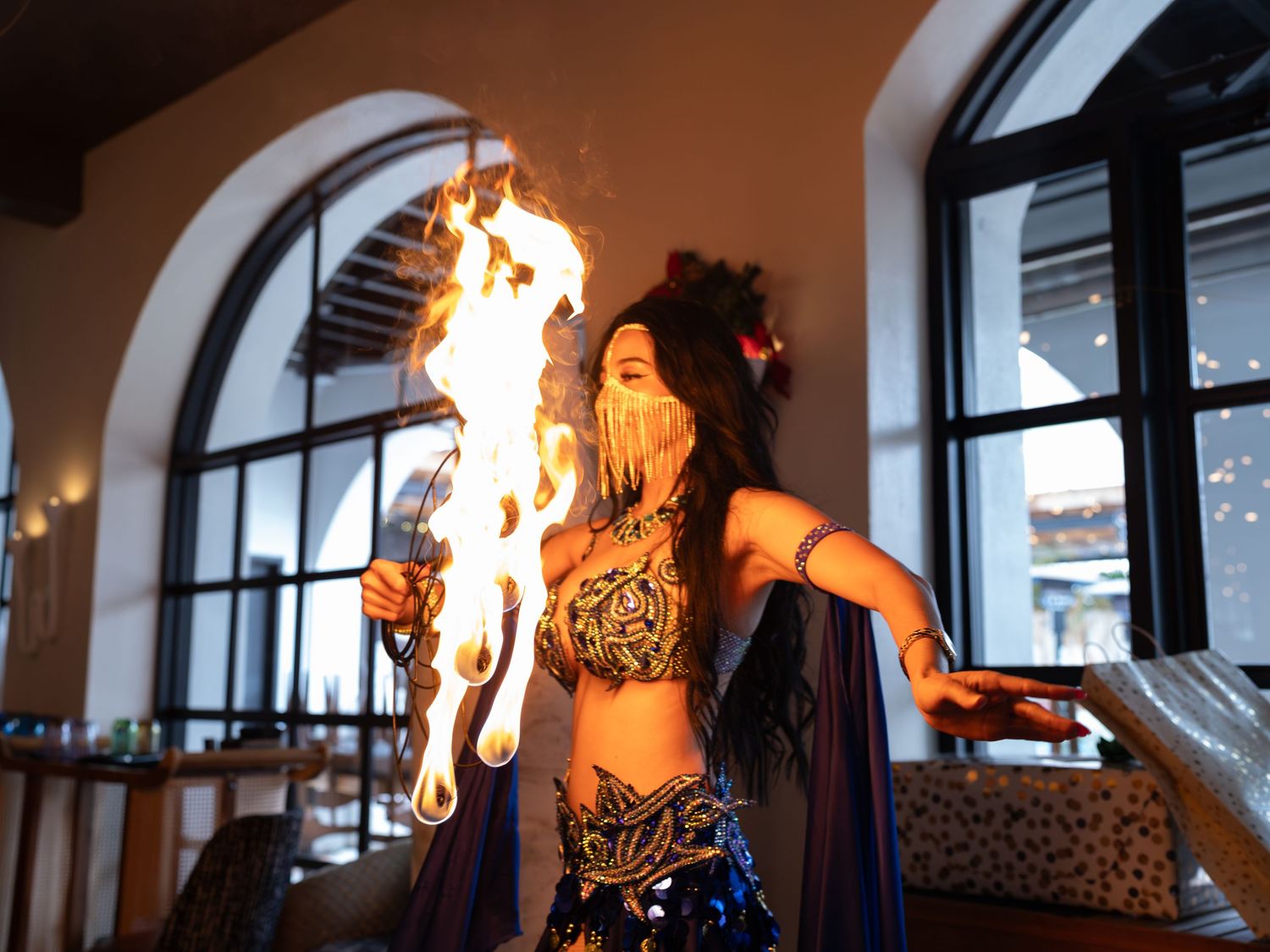 Flames shoot up from a dark interior space with arched windows and dramatic lighting creating a moody atmosphere.