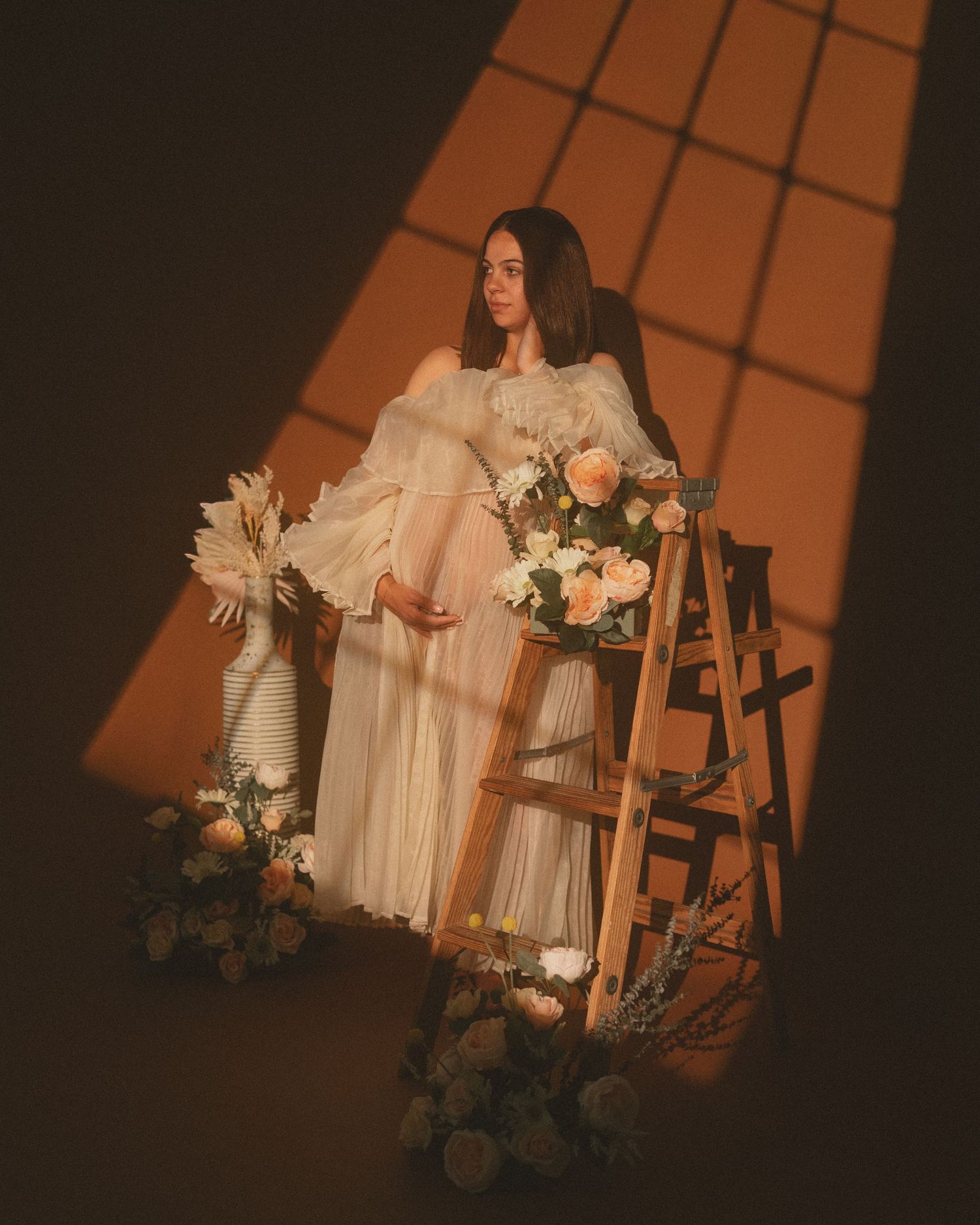 Pregnant woman in white gown sits on wooden ladder surrounded by pastel flowers in warm golden light.