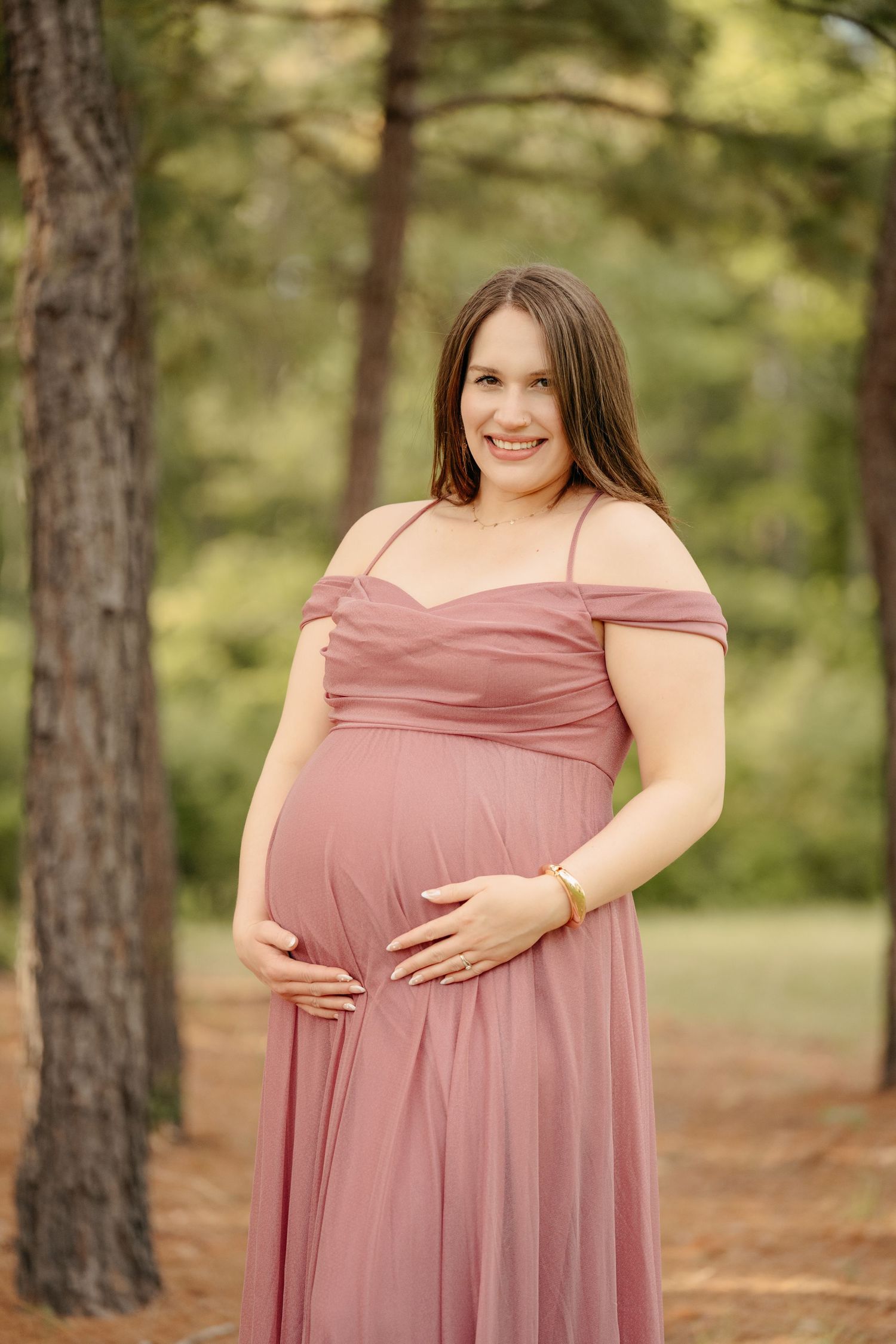 Smiling pregnant woman in a dusty rose off-shoulder gown cradling her baby bump outdoors among pine trees.