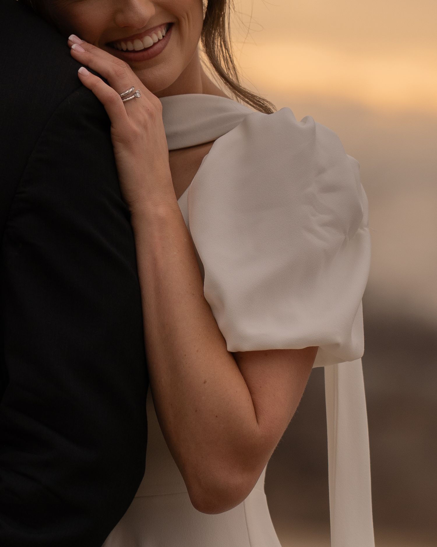 A close-up portrait of someone wearing a white dress with architectural details against a warm sunset background.