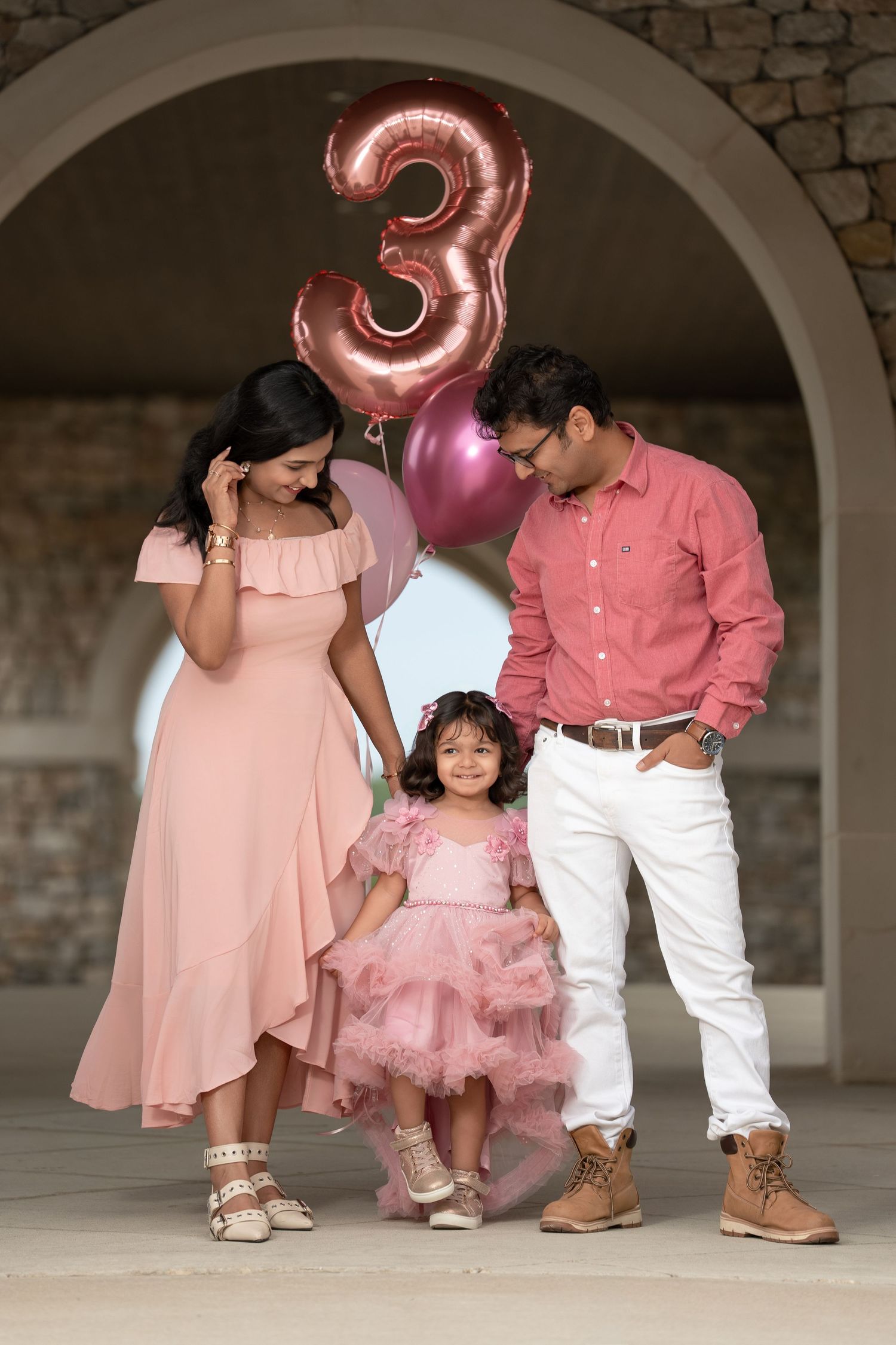 Family of three celebrating a 3rd birthday with pink balloons, wearing coordinating pink and white outfits.