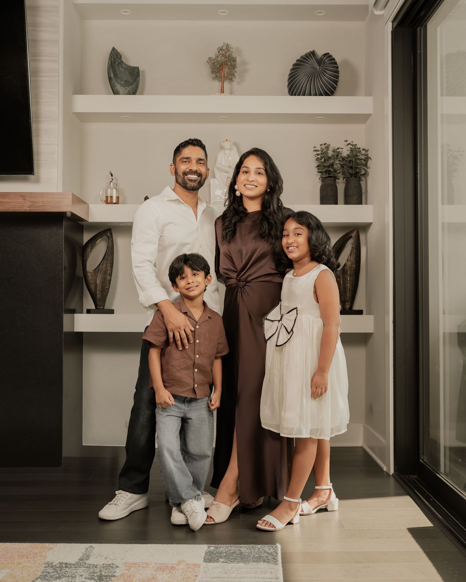 A happy family of four poses indoors, with parents and two children smiling in coordinated brown and white outfits.