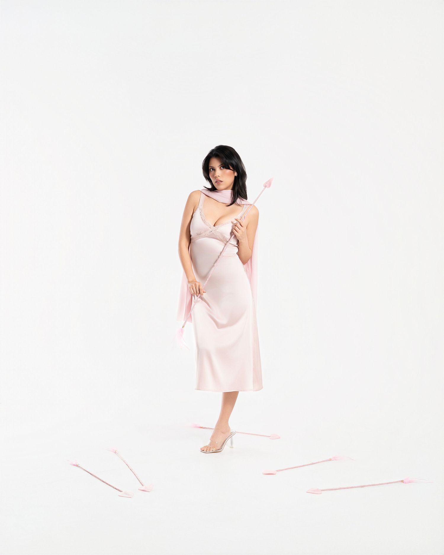 Woman in pink satin slip dress holding Cupid arrows on white background, Valentine's Day fashion editorial.