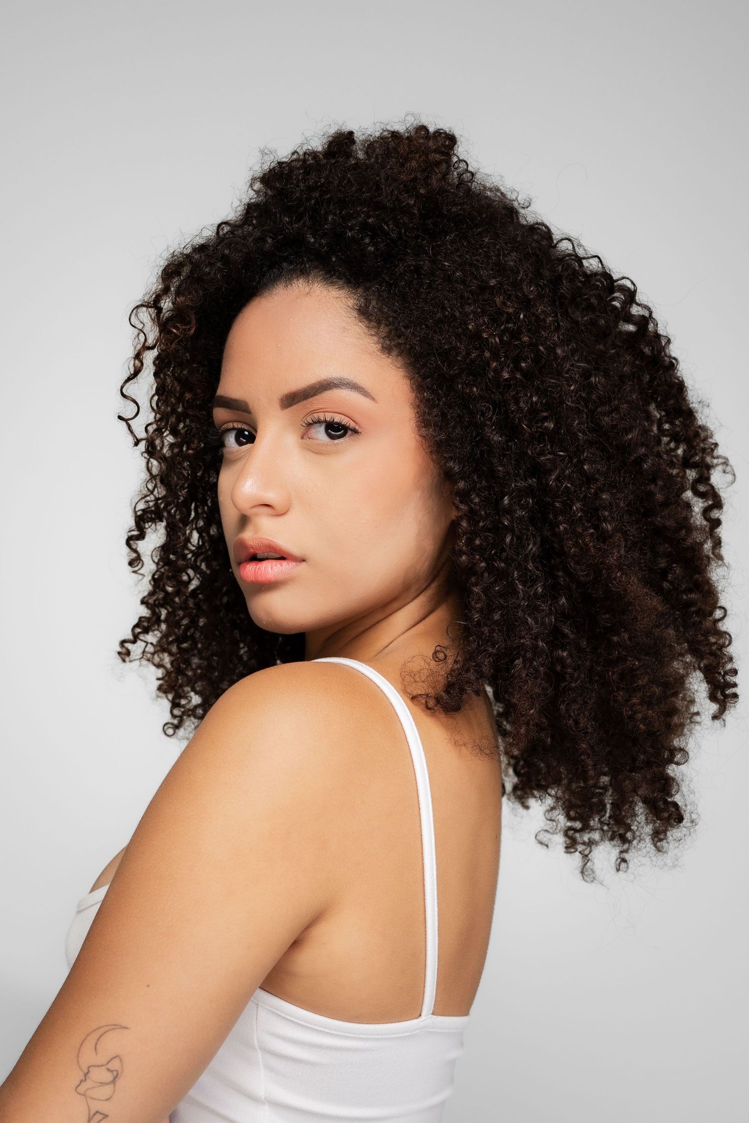 Young woman with voluminous curly dark hair wearing a white tank top, looking over her shoulder against a gray background.