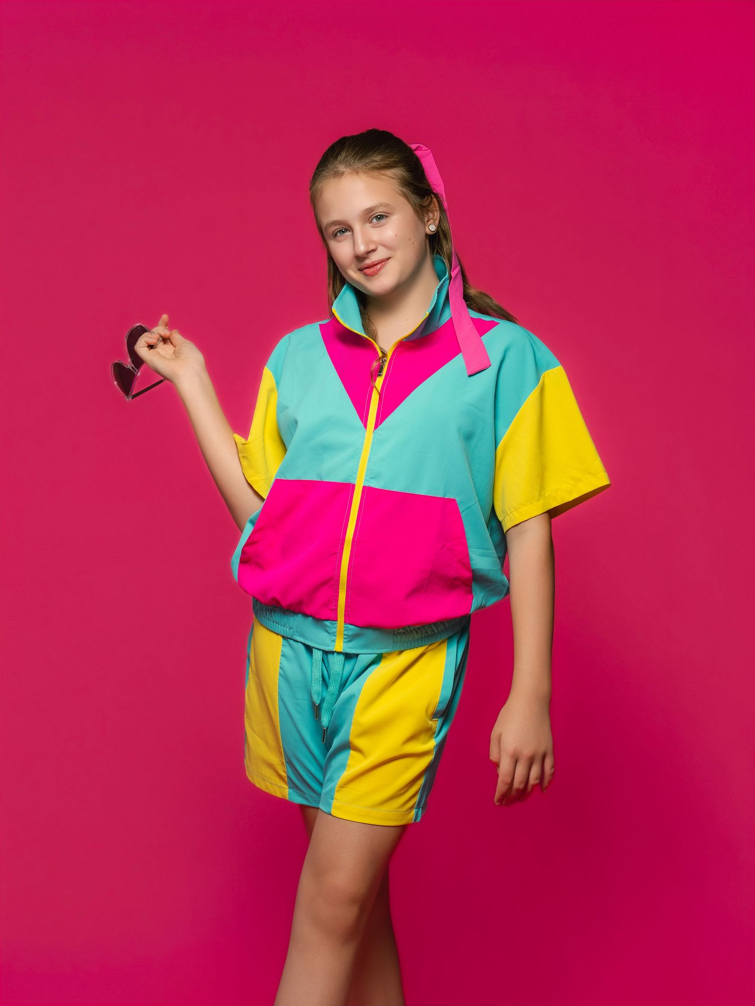 Young girl in colorful 80s-style tracksuit with pink, teal, and yellow colors posing against pink background.
