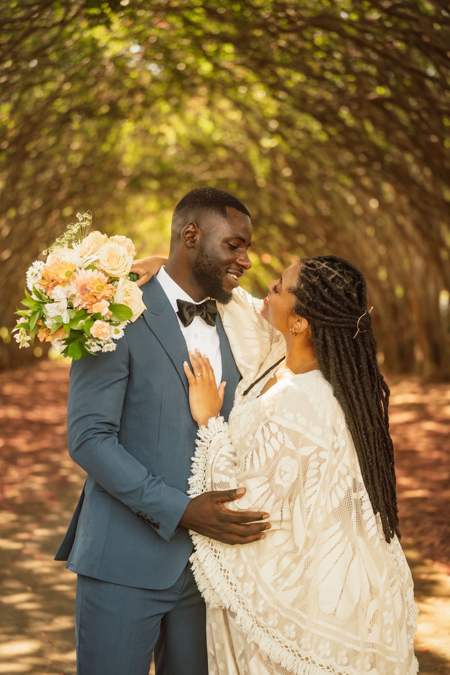 A joyful couple in wedding attire share a romantic moment outdoors under a canopy of trees.
