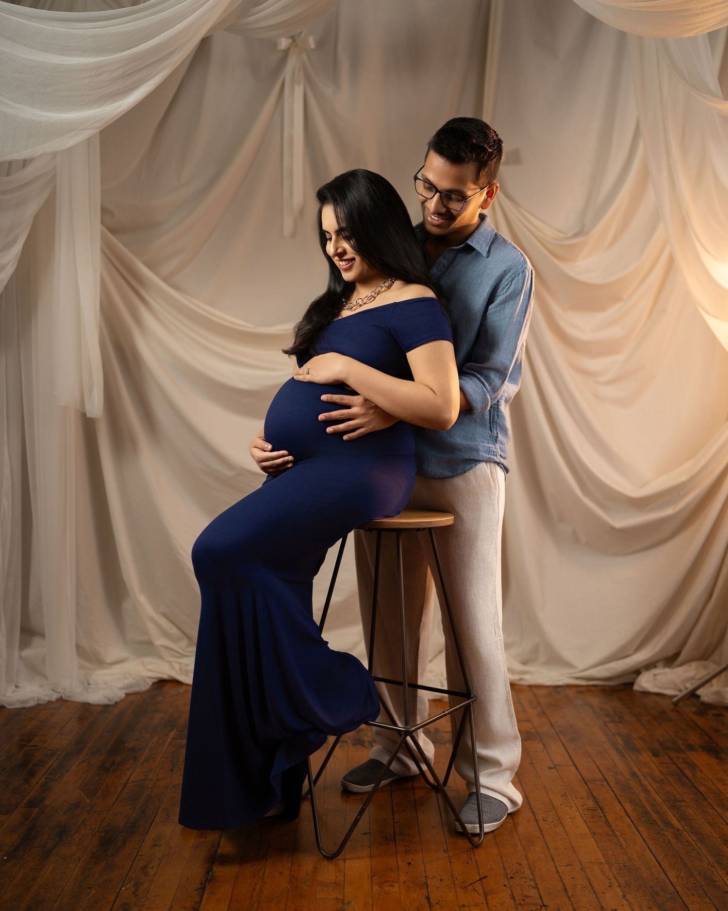 Couple in studio maternity photoshoot, woman in navy gown seated on stool, partner embracing her from behind near white drapes.