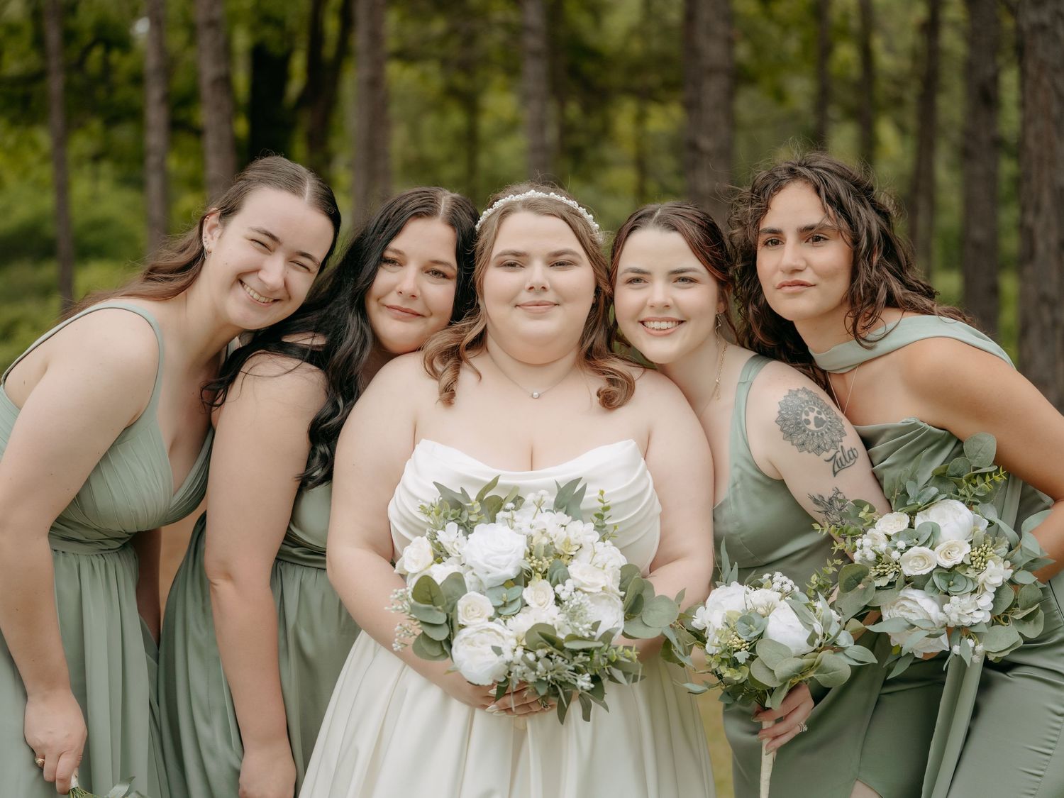 Bride in white gown holding bouquet surrounded by four bridesmaids in sage green dresses outdoors.