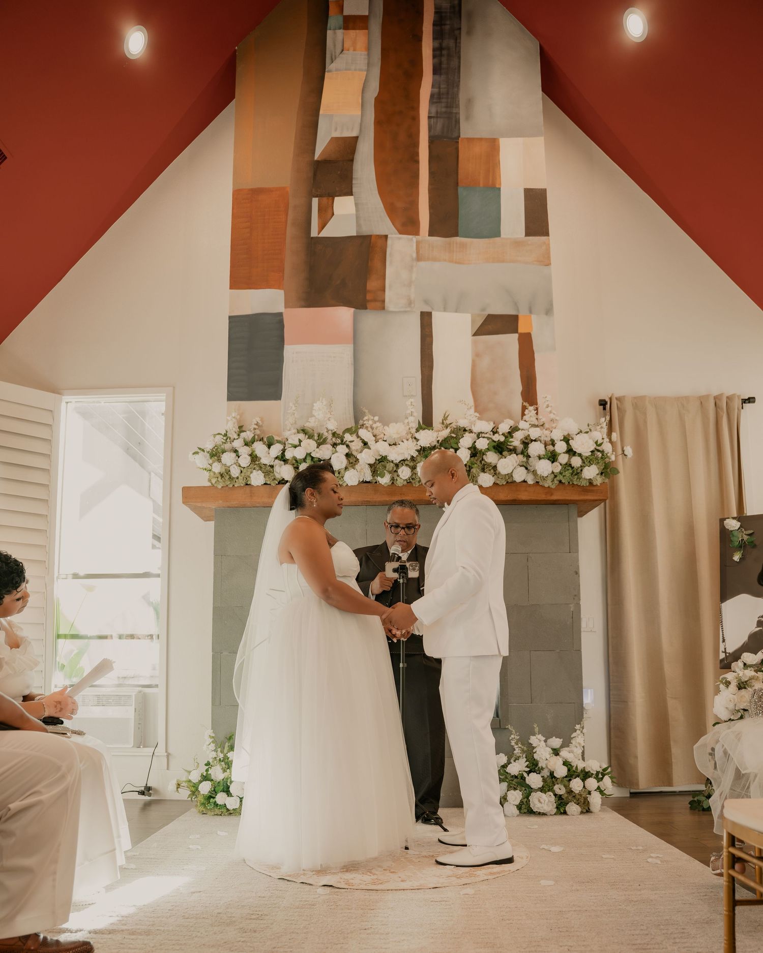 Bride and groom exchange vows at indoor wedding ceremony with white floral arrangements and colorful abstract art backdrop.