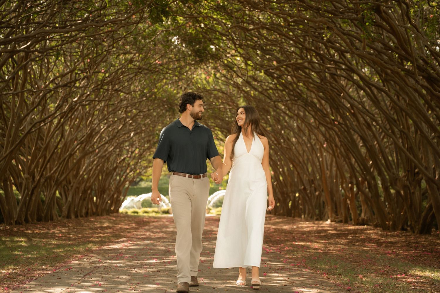 Couple holding hands walking under a natural tree tunnel archway on a scenic garden path.