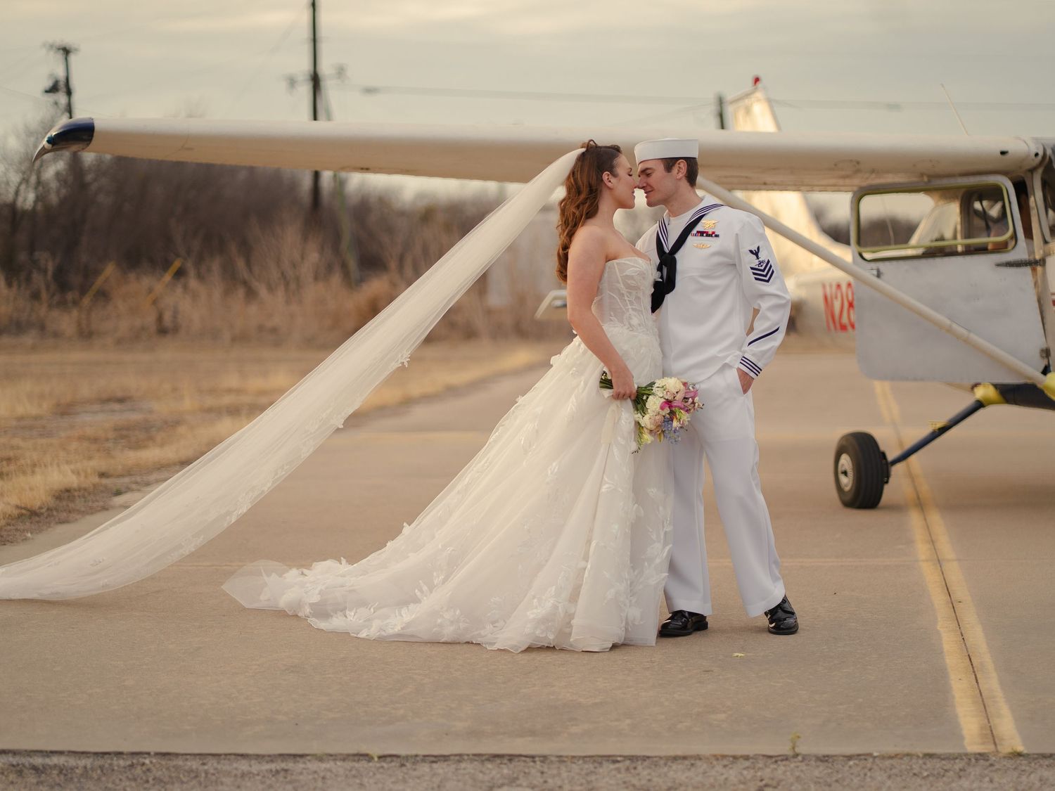 A couple in wedding attire poses romantically on an empty road at sunset with the bride's dress flowing in the wind.