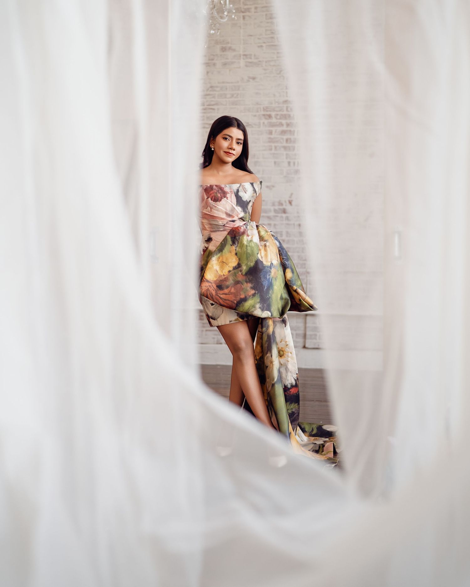 Woman in colorful floral off-shoulder dress poses elegantly between sheer white curtains in a bright studio.