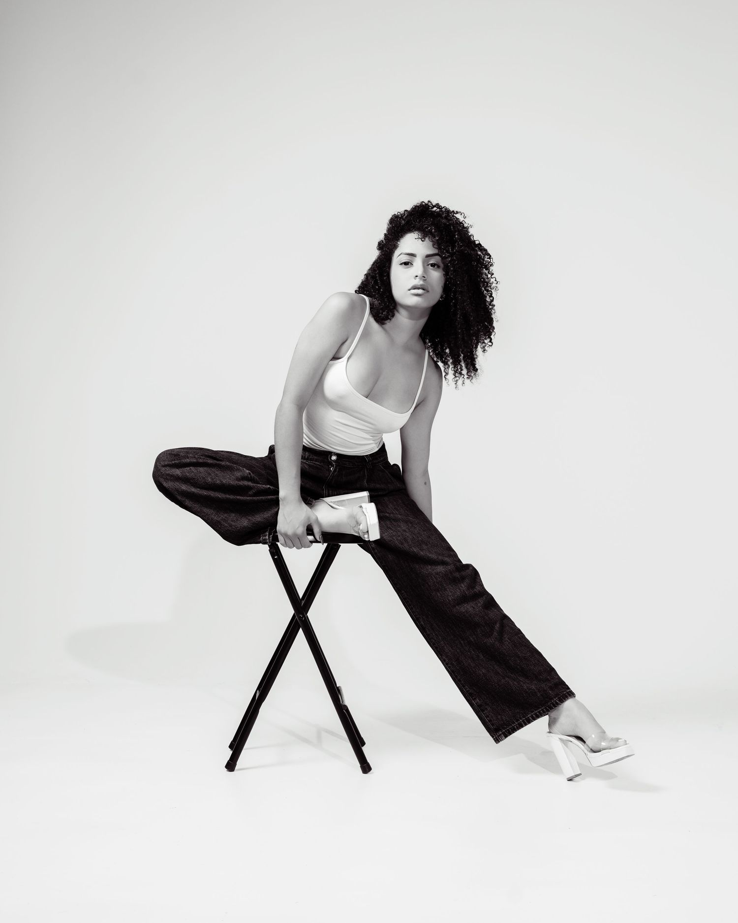 Black and white fashion photo of a woman in wide-leg pants and white bodysuit posing dynamically on a stool.