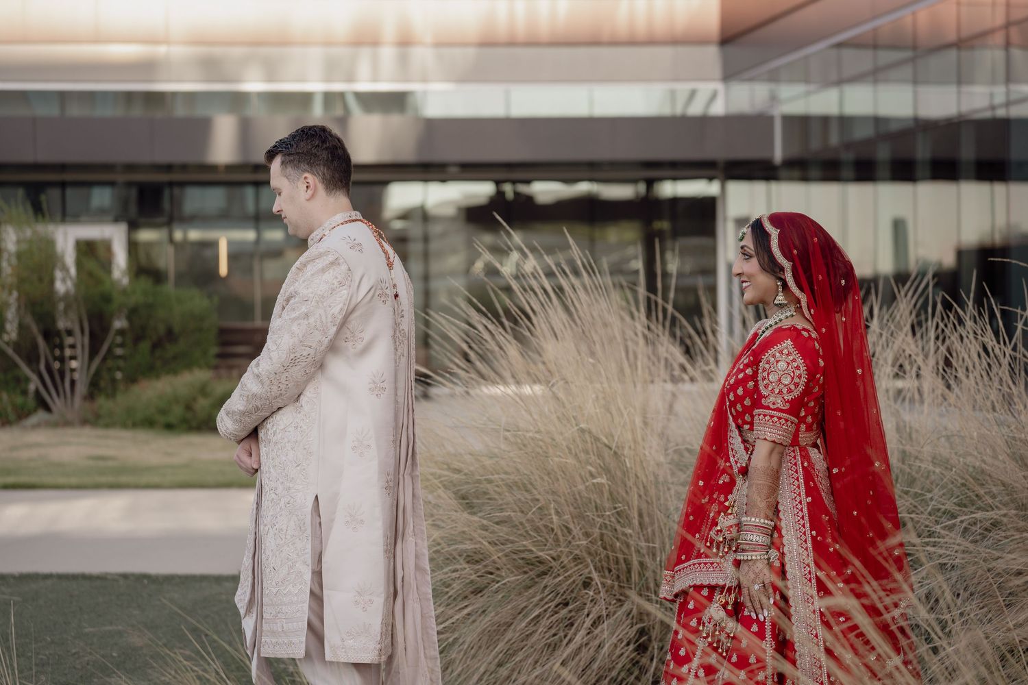 Indian bride in red lehenga and groom in cream sherwani share a first look moment outdoors at a South Asian wedding.